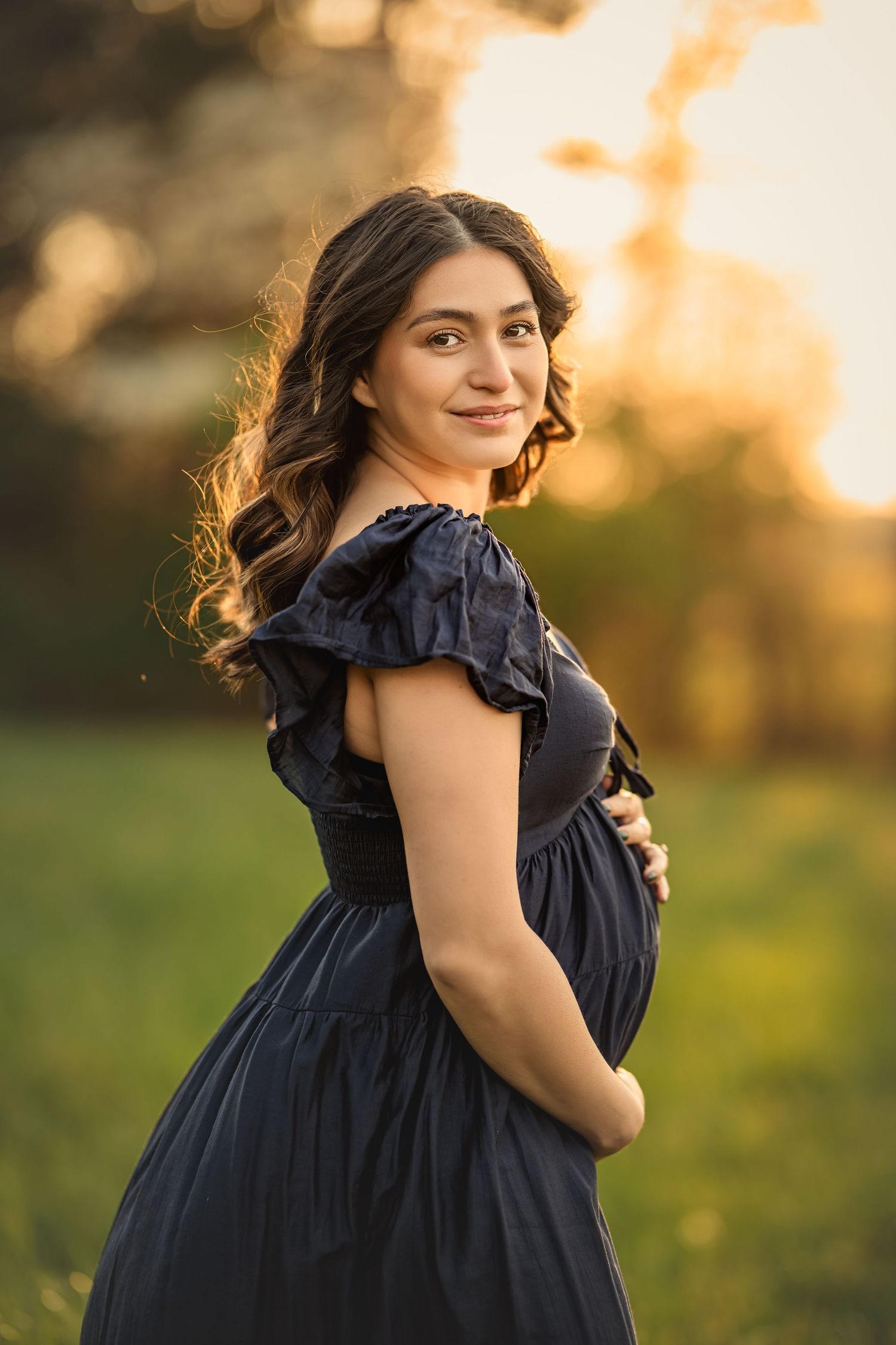 Pregnant woman in navy blue dress holding small baby bump at golden hour sunset in a green field for atlanta maternity photos