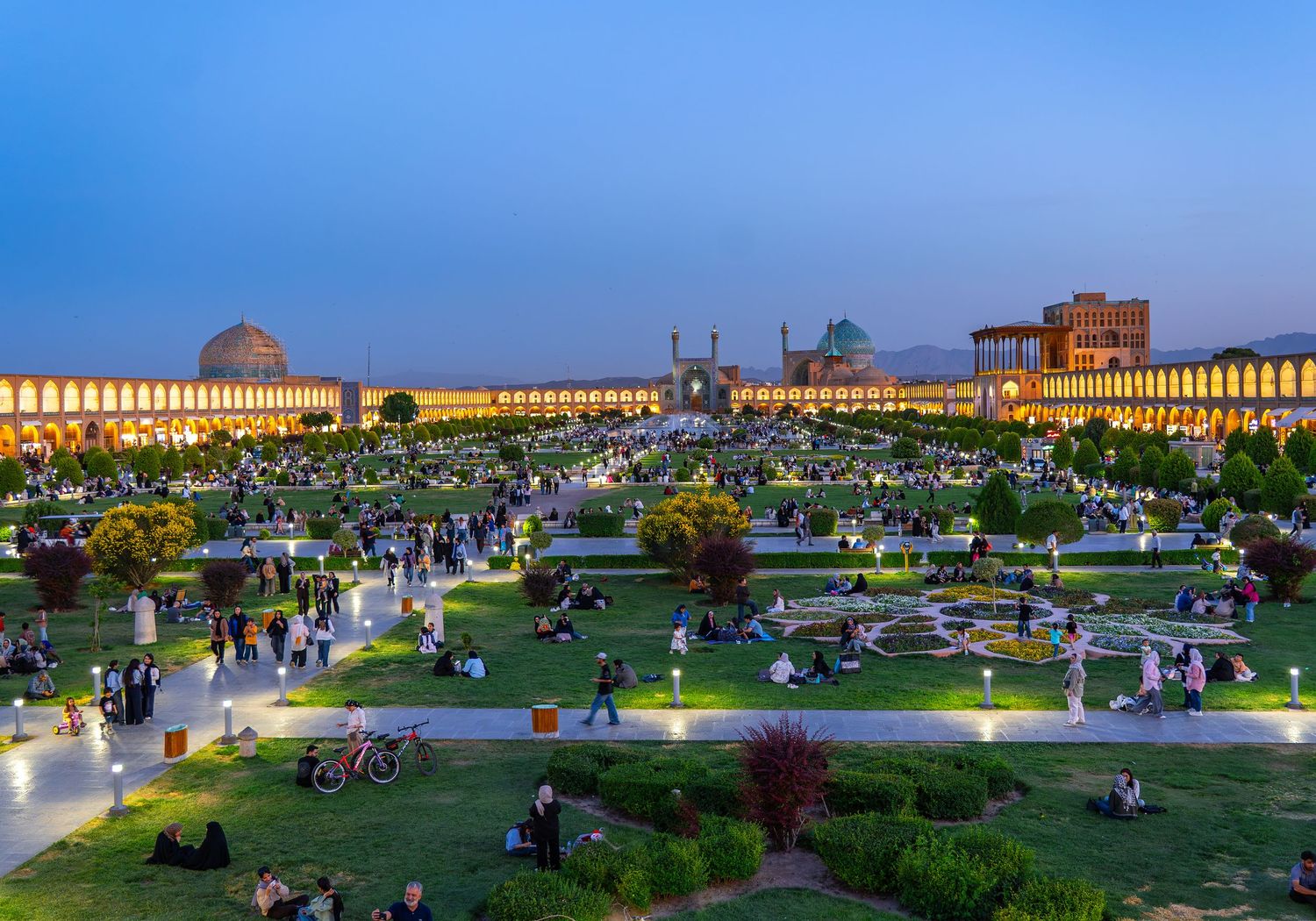 Naqsh-e Jahan Square in Isfahan, Iran, a UNESCO World Heritage Site, at dusk with illuminated mosque domes and people gathered on the lawn.