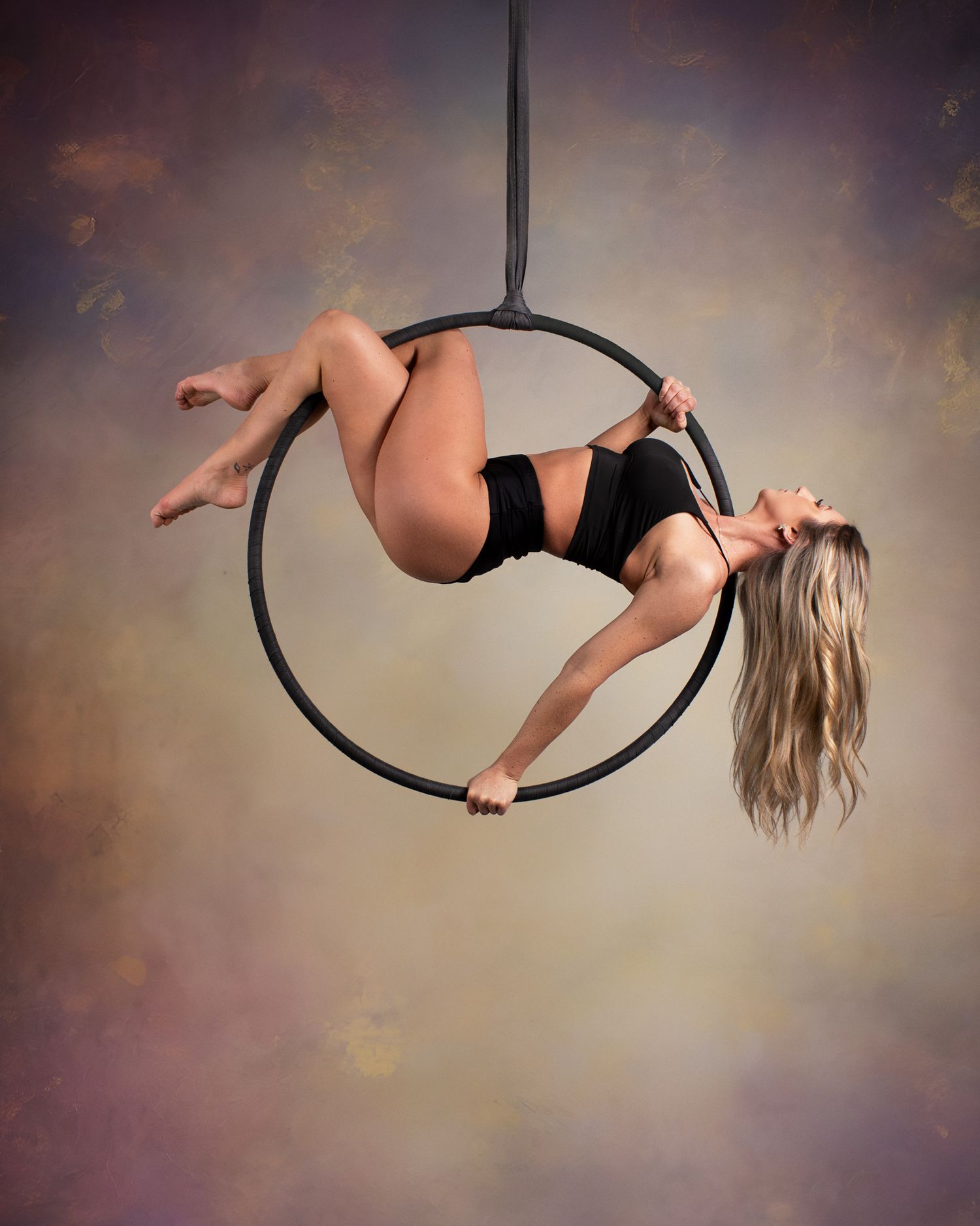 Aerial hoop performer in black outfit gracefully balancing on a lyra ring suspended mid-air against colorful backdrop.