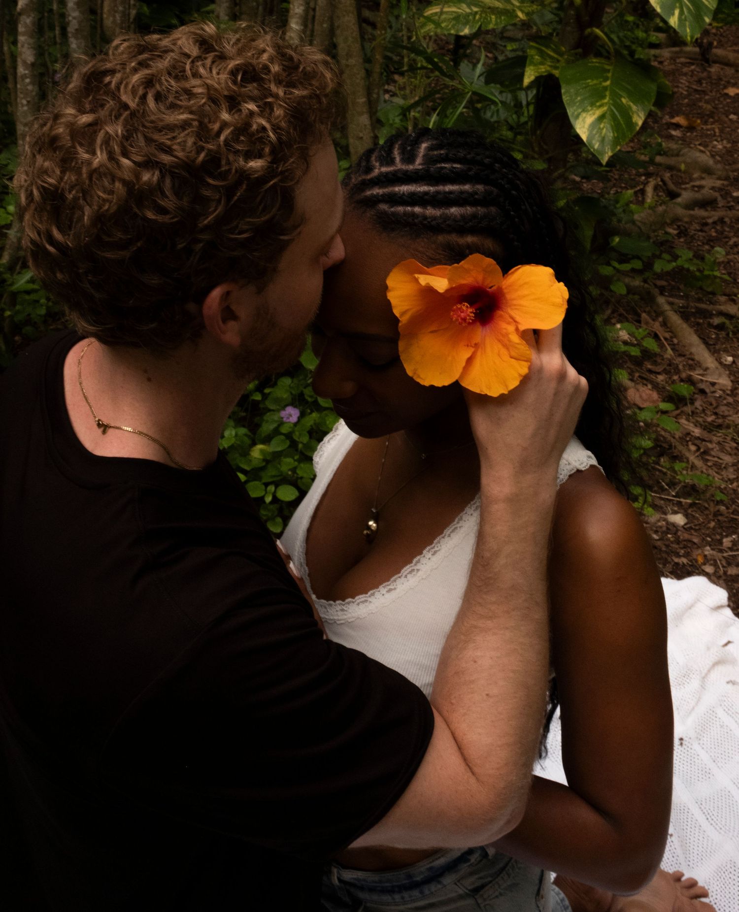 Couple sharing a romantic moment outdoors, woman with braids holding an orange hibiscus flower, surrounded by lush greenery on Oahu, Hawaii.