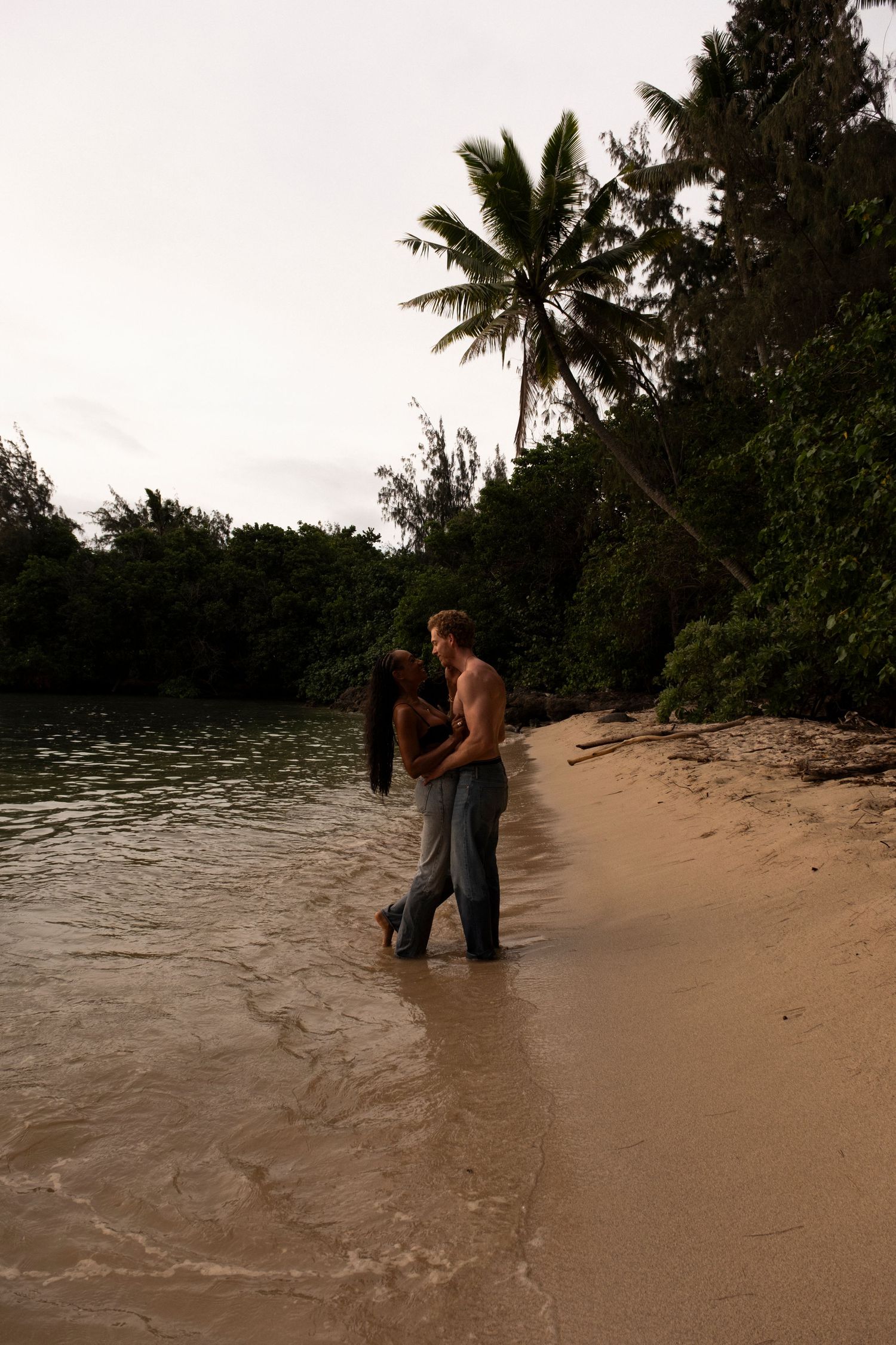 Romantic couple embracing on a tropical beach shore with palm trees and lush greenery at dusk on Oahu, Hawaii.