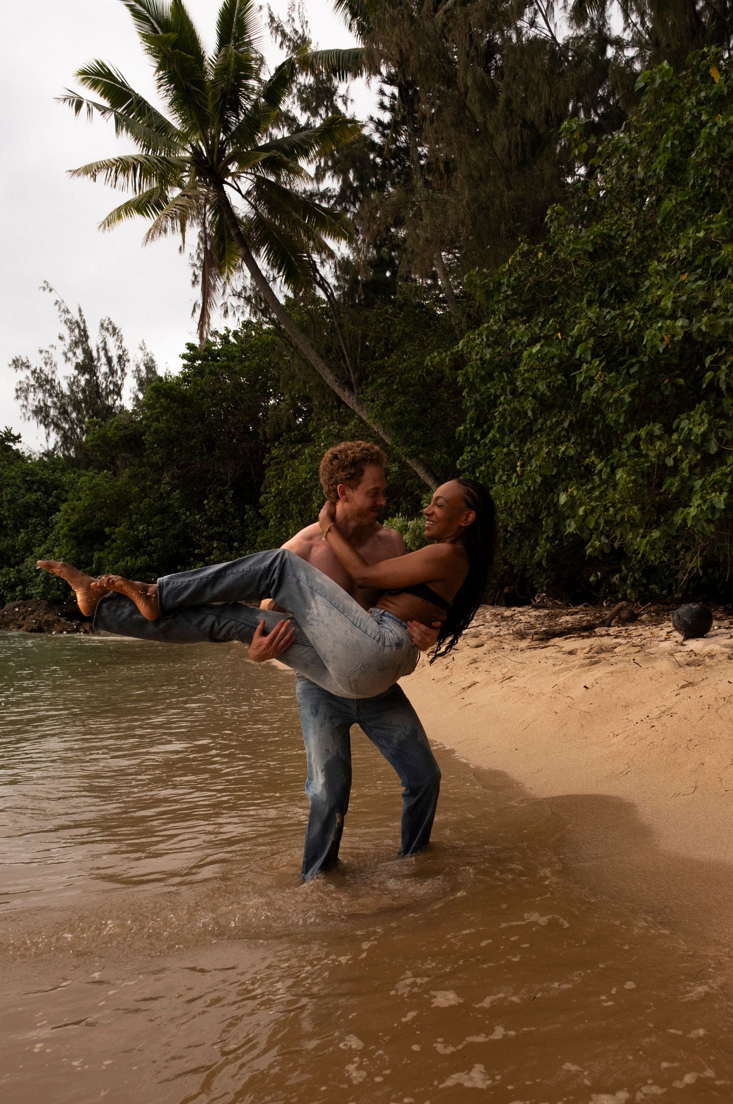 Couple embracing romantically on a tropical beach shore, man carrying woman in his arms near palm trees and calm water on Oahu, Hawaii.