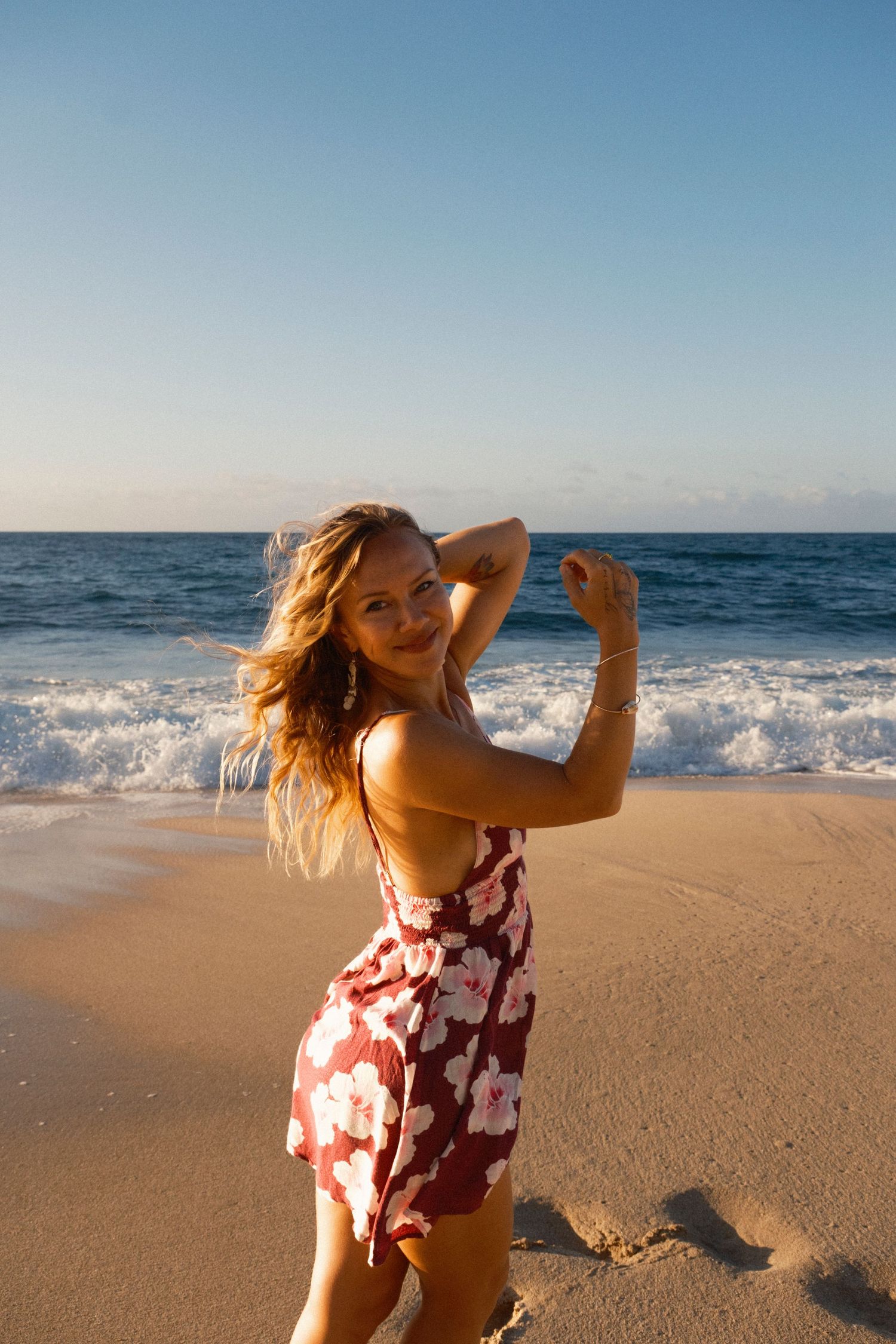 Young woman in red floral dress posing on sandy beach with ocean waves at golden hour sunset on Oahu, Hawaii.