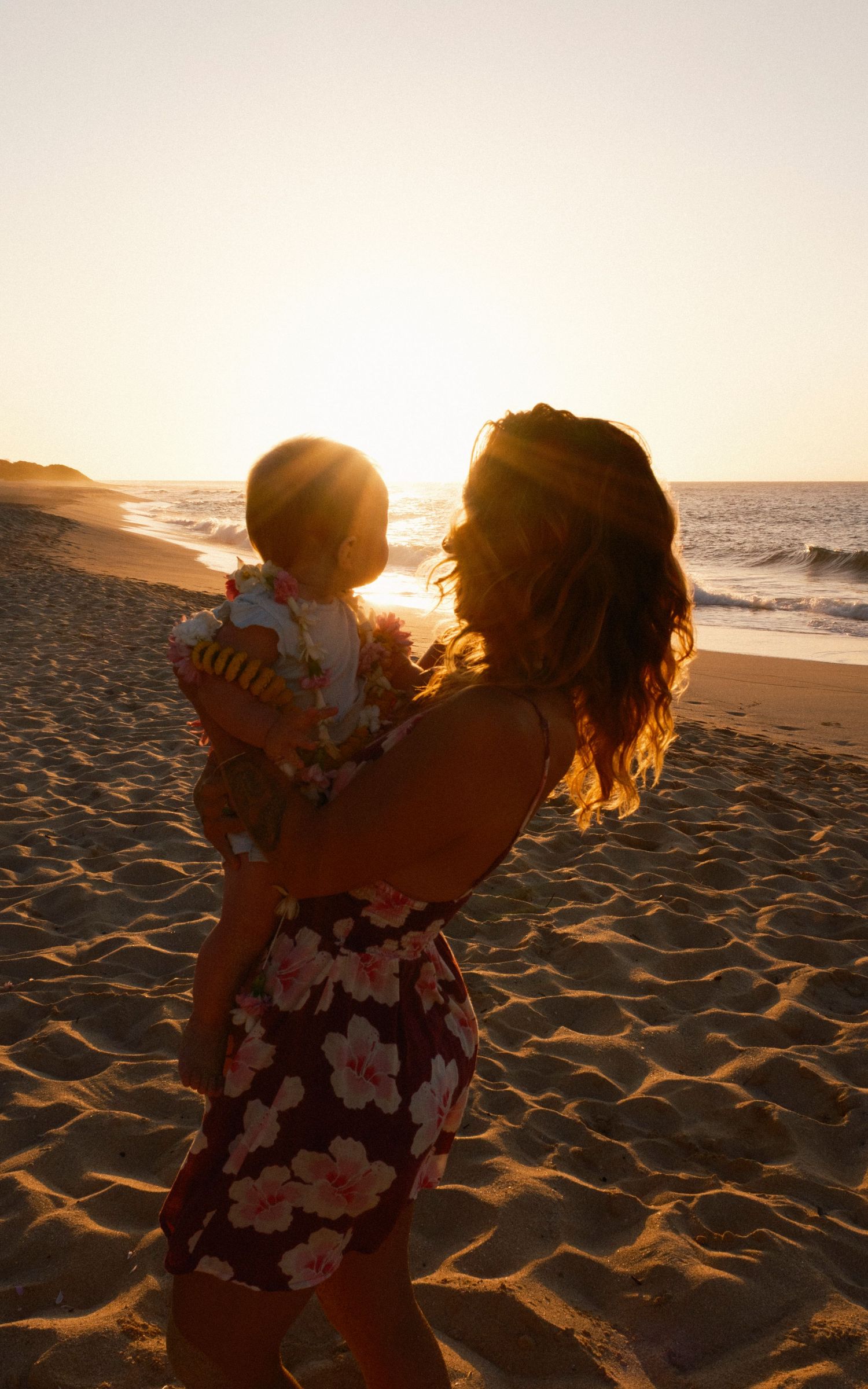 A mother holds her baby on a sandy beach at golden sunset, both silhouetted against the glowing ocean horizon on on Oahu, Hawaii.
