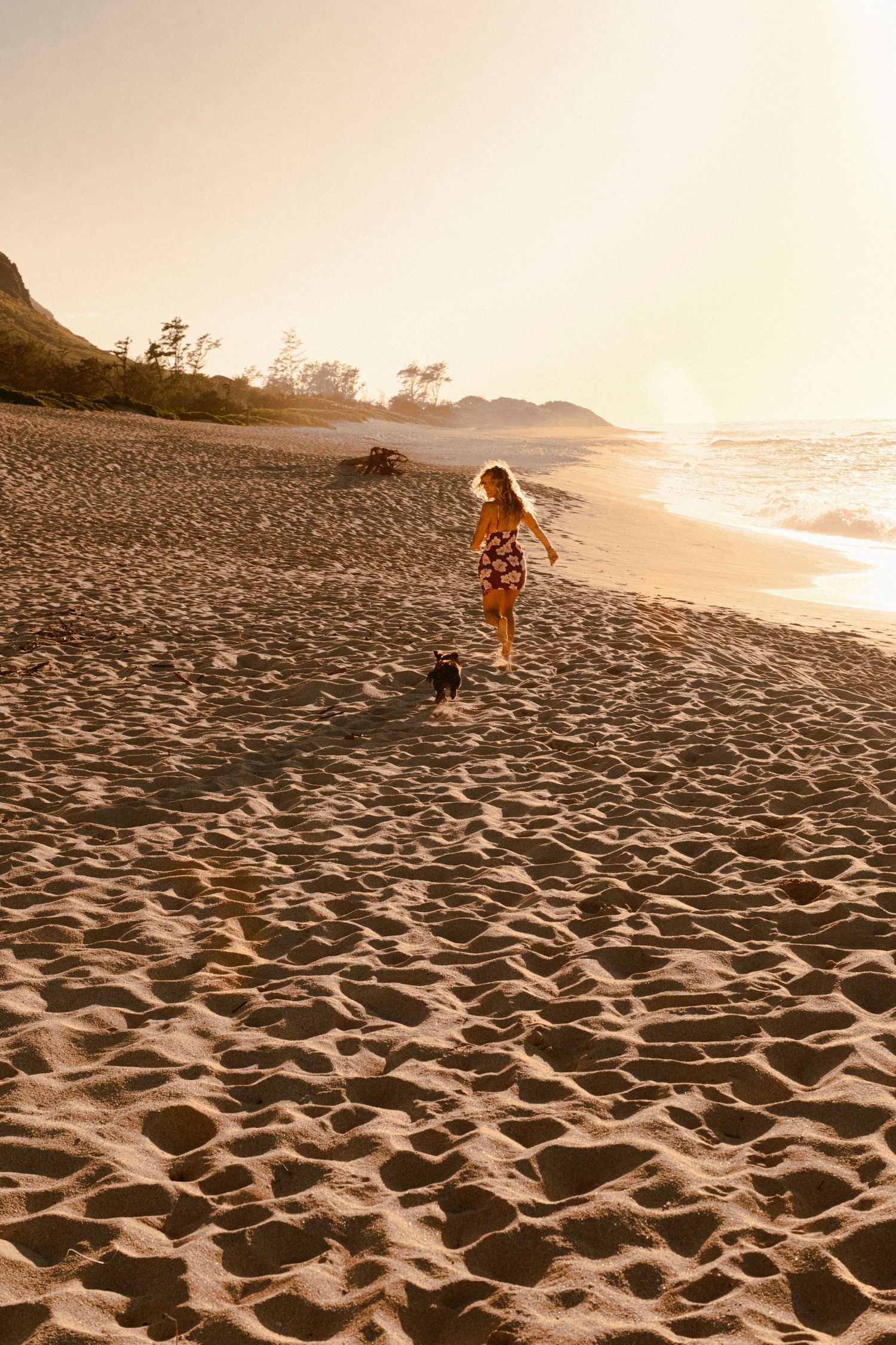 Woman in floral dress running with small dog on sandy beach at golden sunset hour on Oahu, Hawaii.