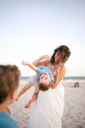 Laughing mother playfully holds child upside down on a sunny beach while another child watches nearby.