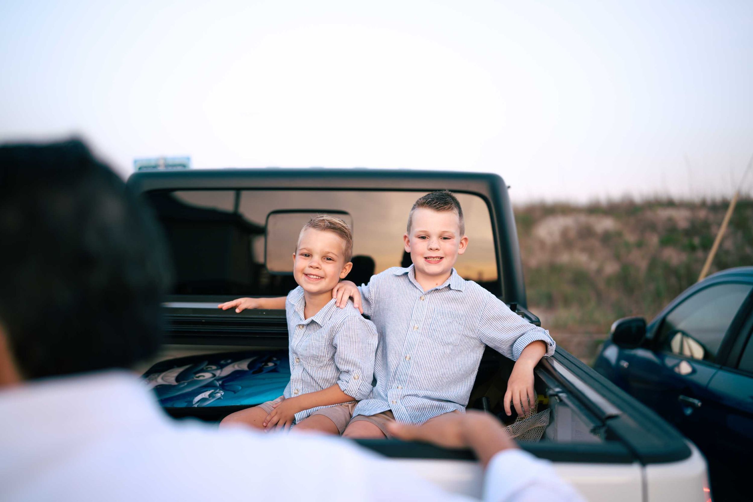 Two young brothers in matching striped shirts sit together in the back of a pickup truck near the beach.