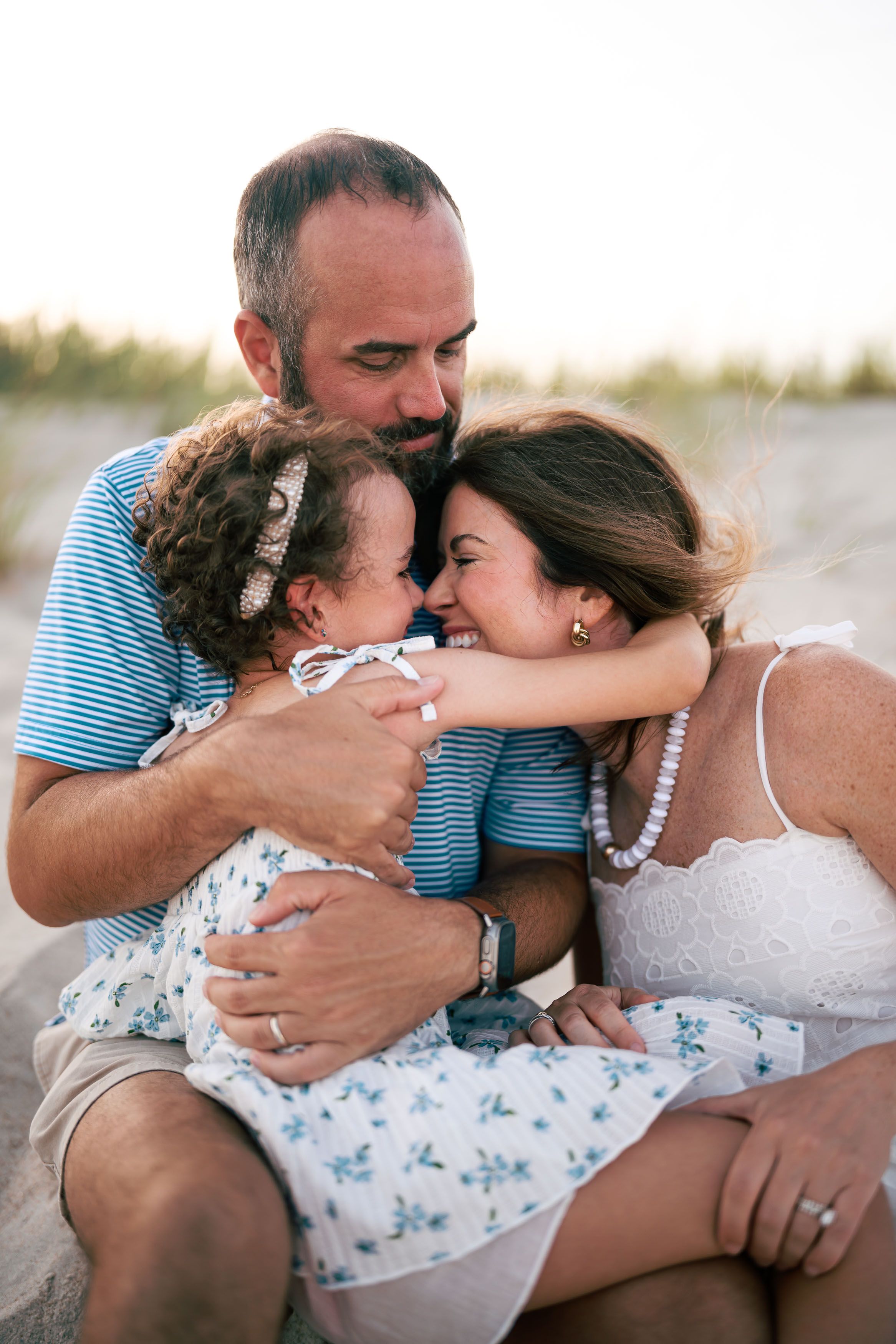 Family of three sharing a warm embrace on the beach at sunset, parents hugging their young daughter.