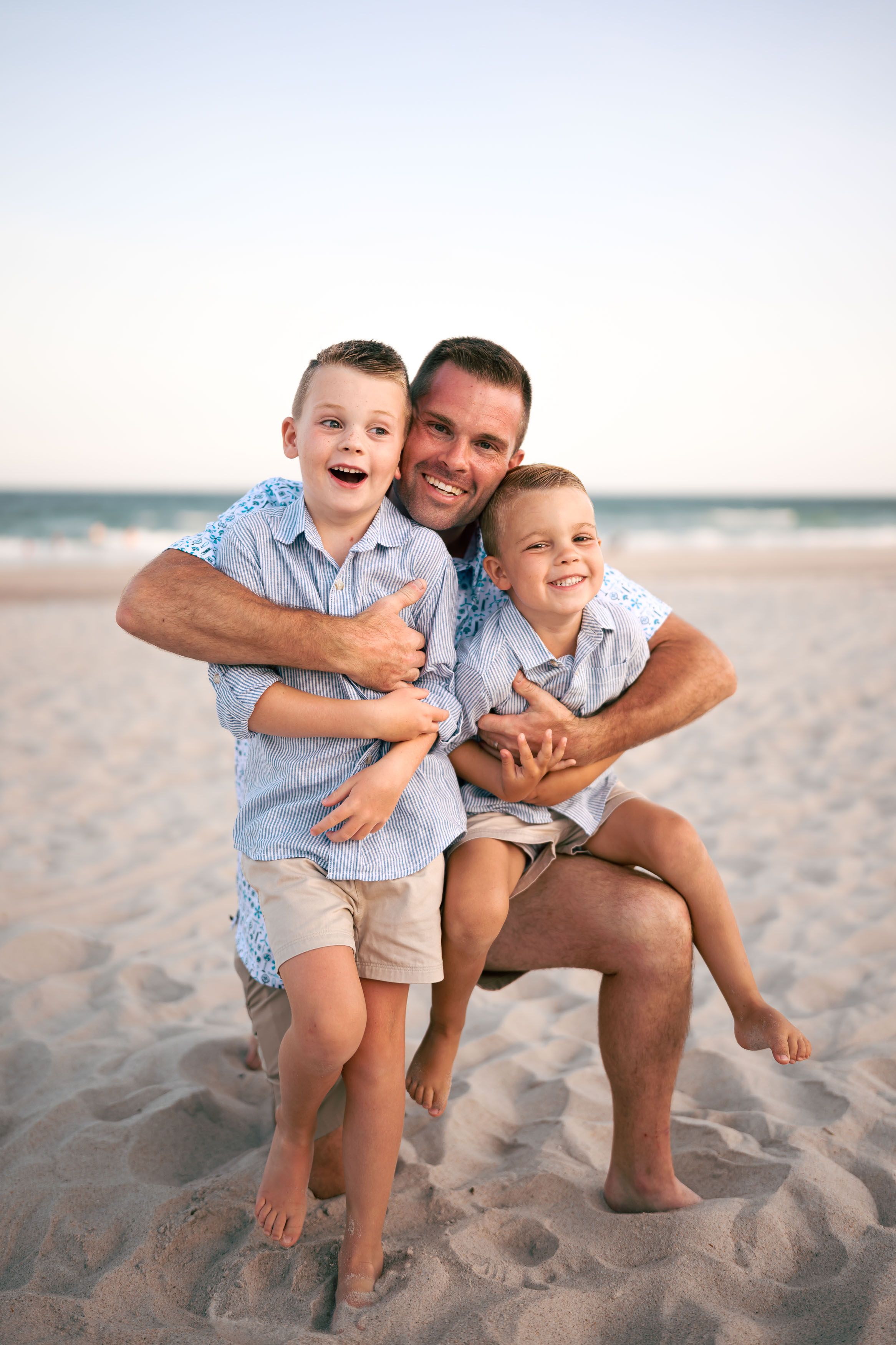 A smiling father hugs his two young sons on a sandy beach at sunset, all wearing matching blue shirts.
