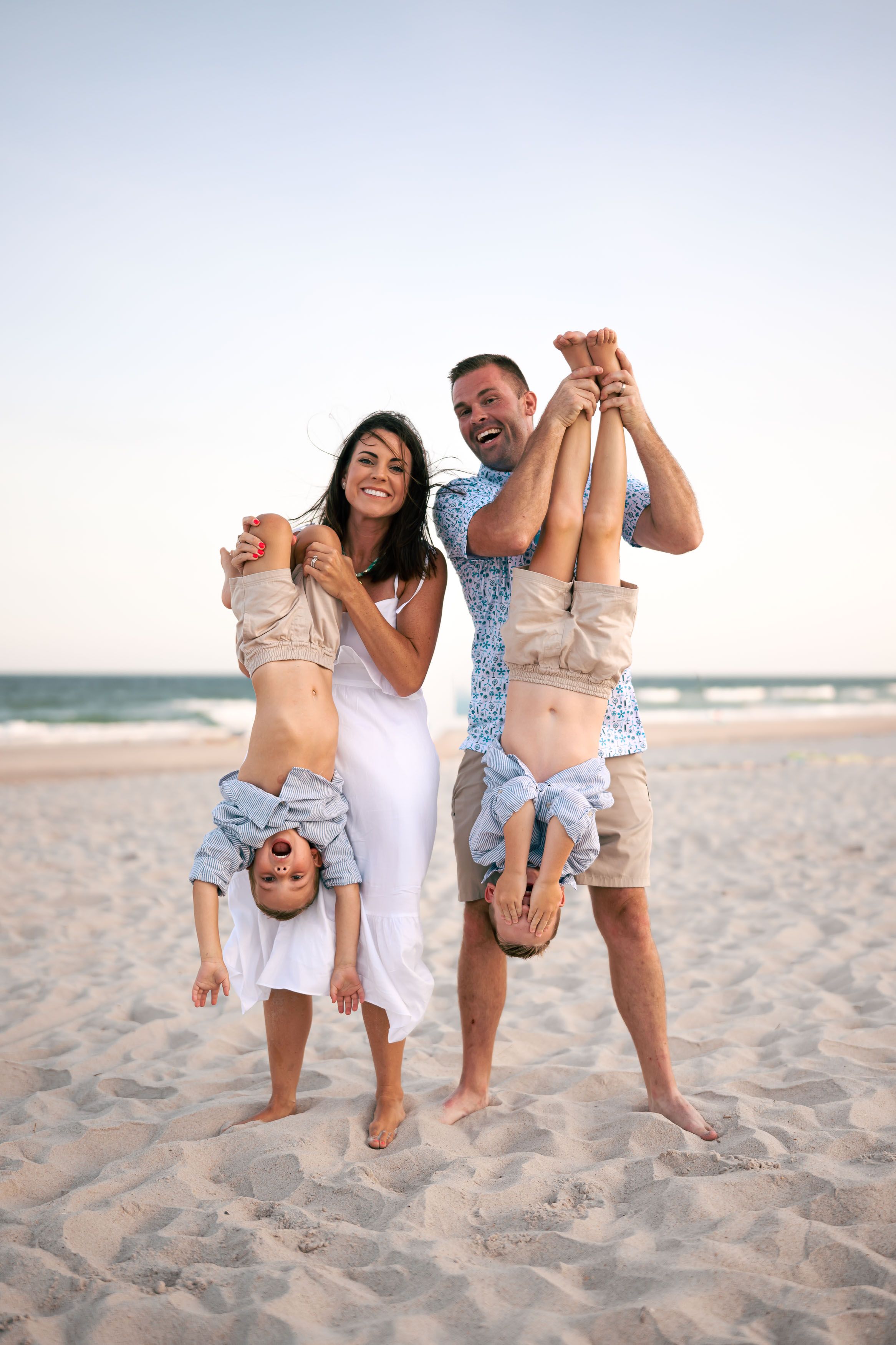 Happy family laughing on beach as parents playfully hold two children upside down on sandy shore.