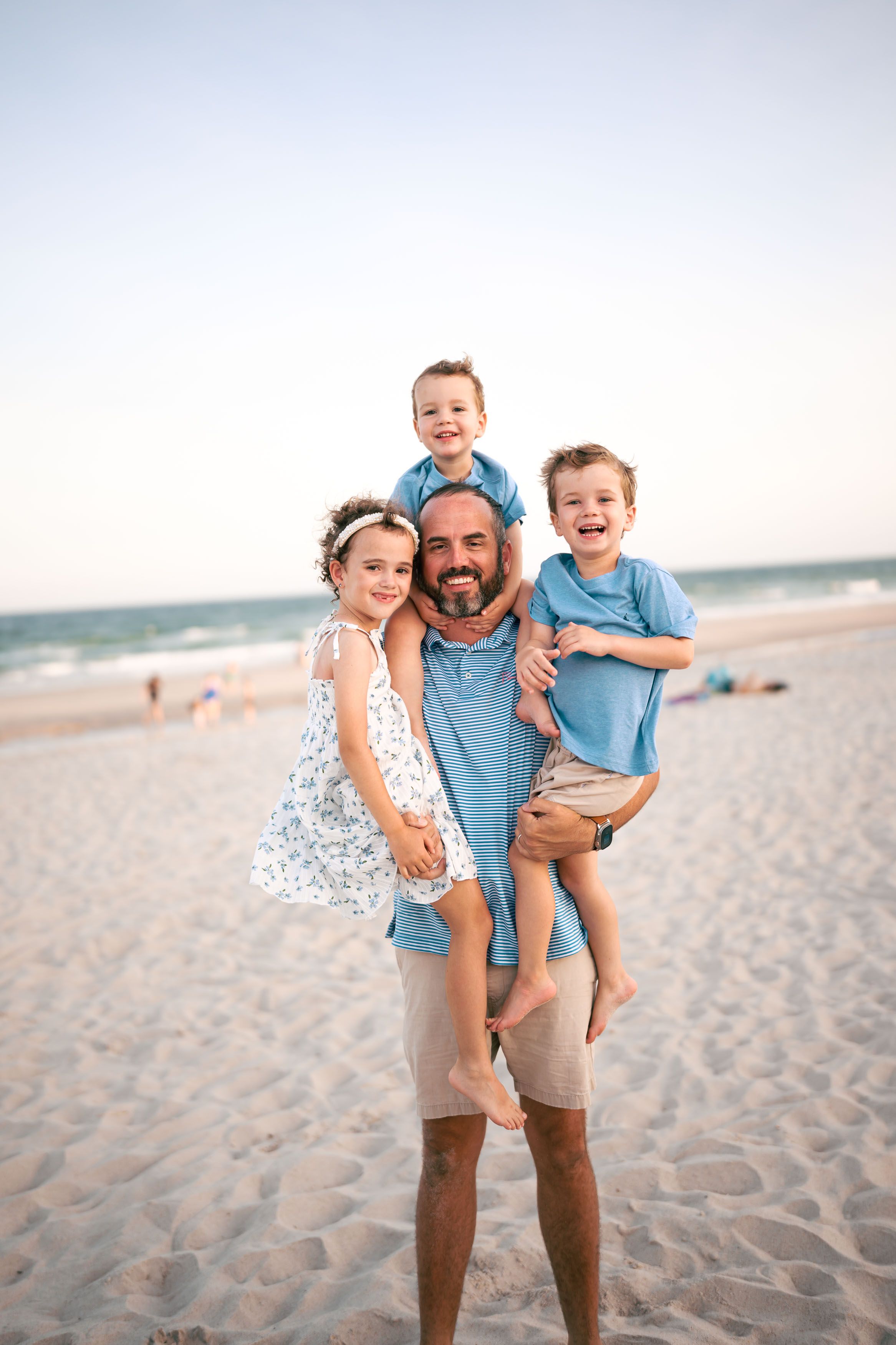 Happy father holds three young children on a sandy beach, all smiling in casual blue and white outfits.
