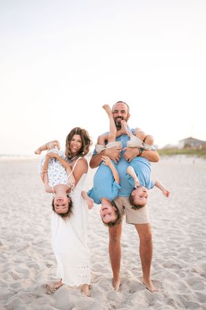 Happy family of five playfully holding children upside down on a sunny beach.
