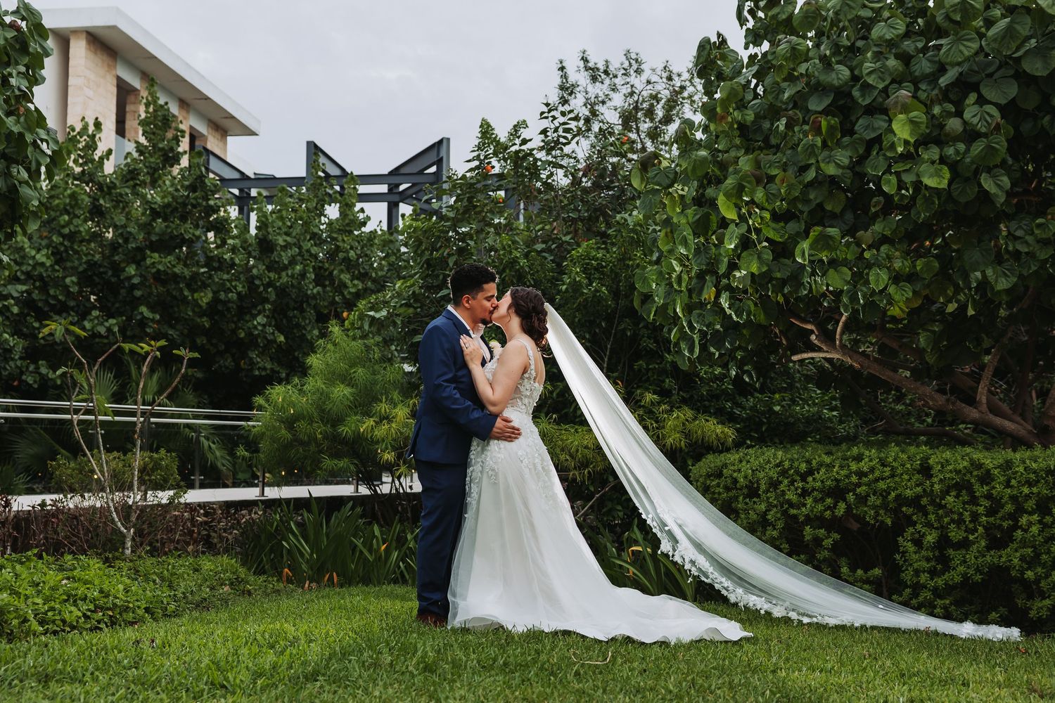 Bride in white strapless gown sits on groom's lap as he kisses her cheek, surrounded by lush garden greenery.