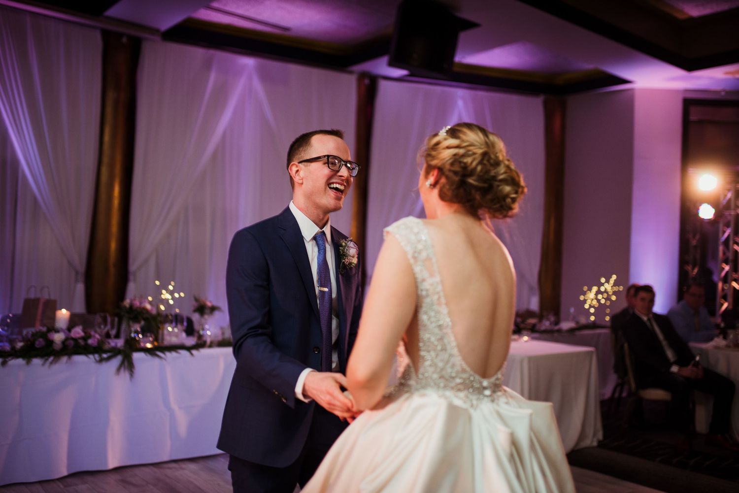 A groom in a navy suit laughs joyfully with his bride in a white gown during their first dance at a wedding reception.
