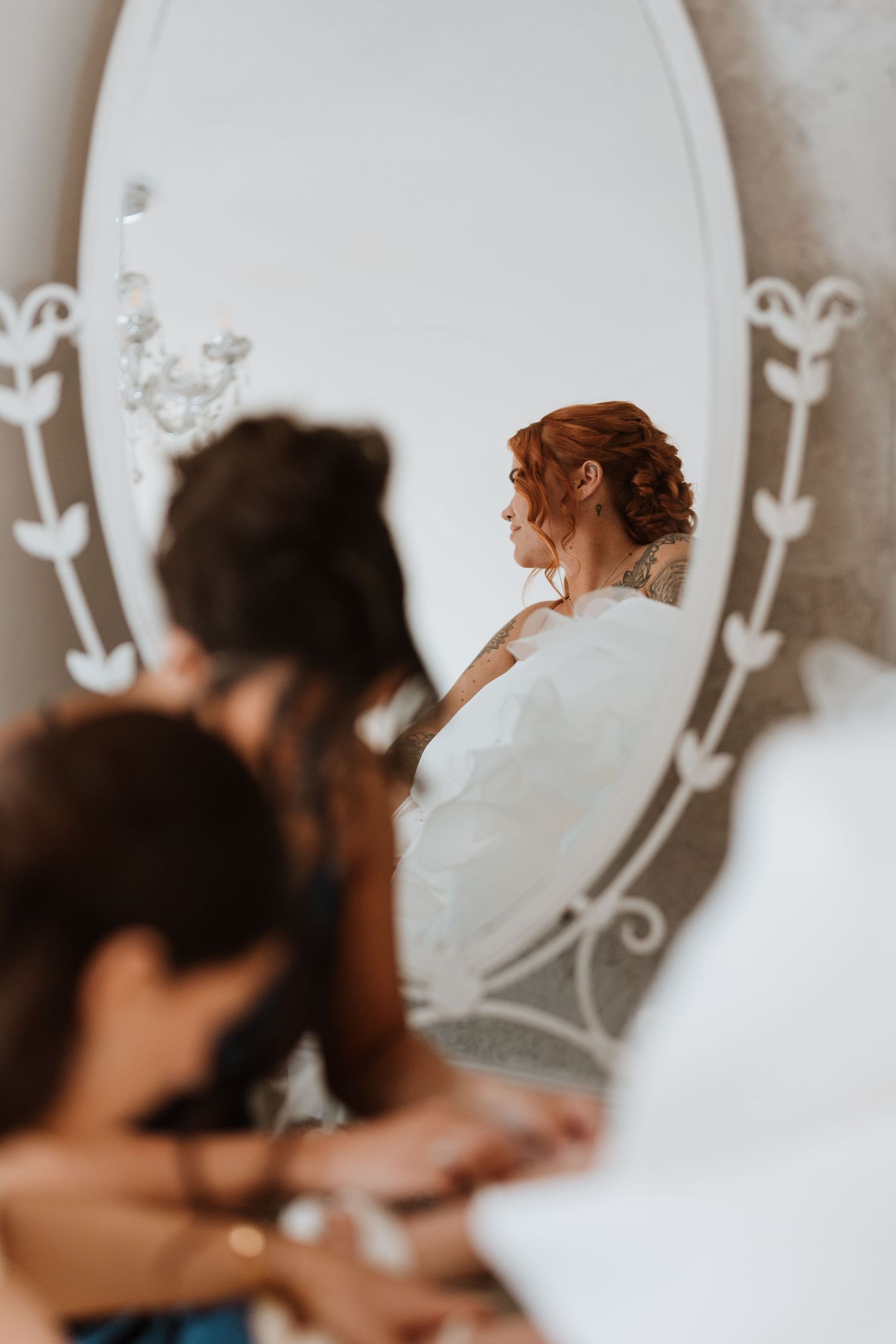 Bride in white dress reflected in ornate oval mirror, with bridesmaids in foreground during wedding preparations.