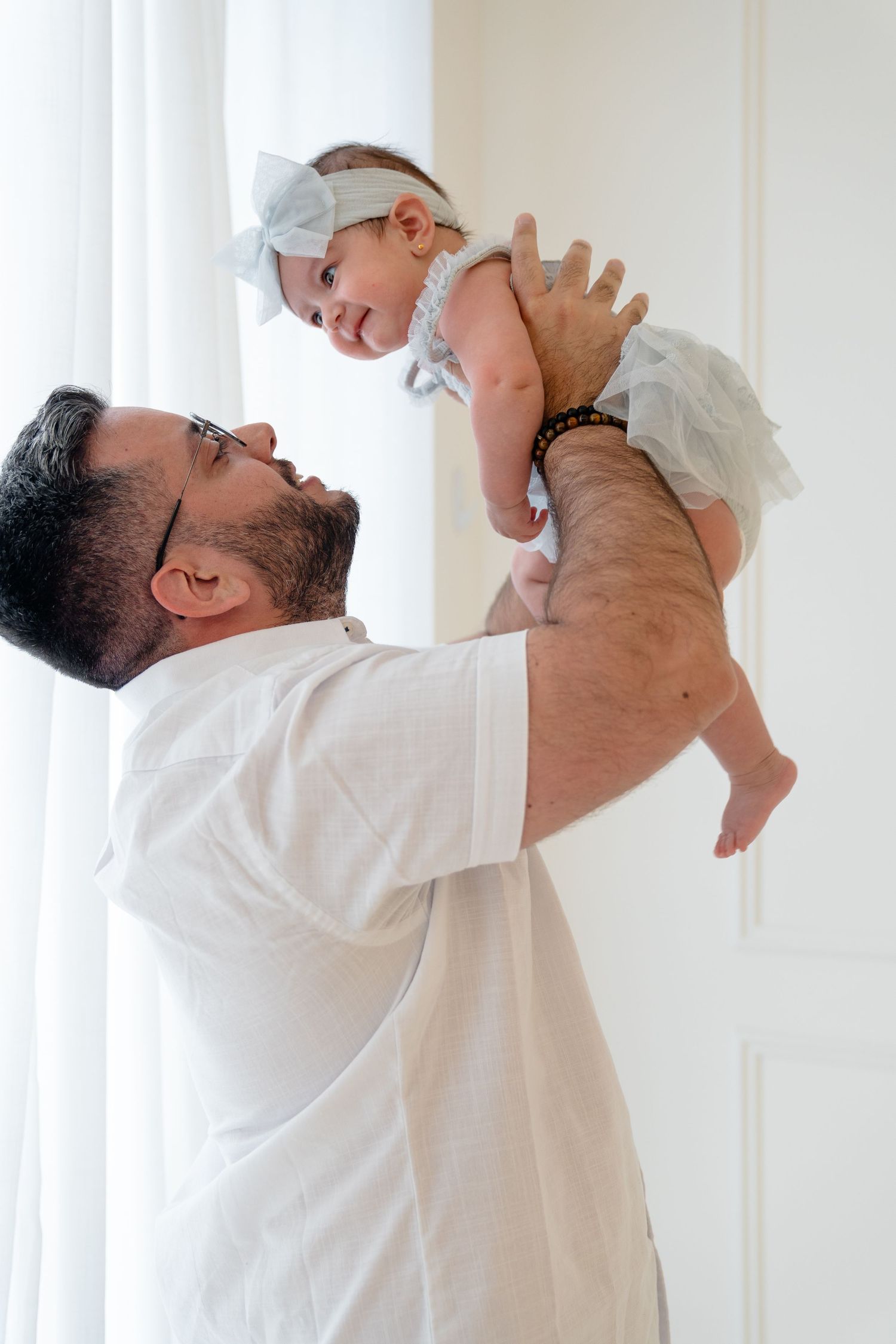 A bearded father in a white shirt joyfully lifts his smiling baby girl dressed in a white tutu near a bright window.