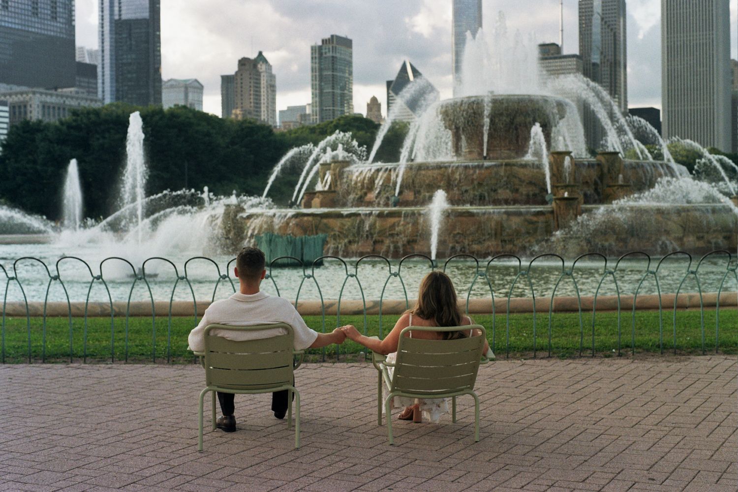Couple sitting on outdoor steps embracing near a fountain with city skyline in background.