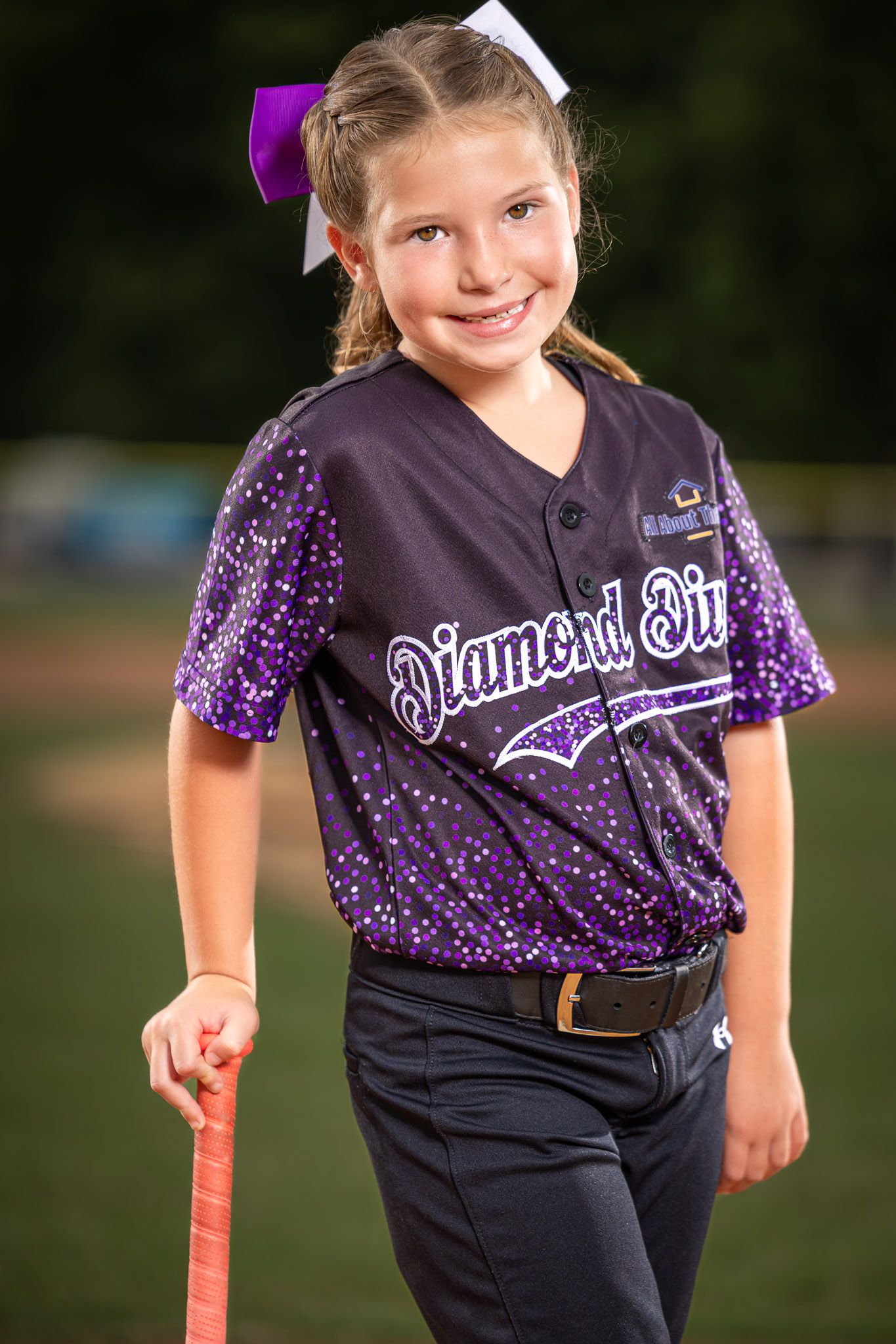 Young girl in Diamond Diva softball jersey holding bat, smiling on field wearing purple bow and navy pants.