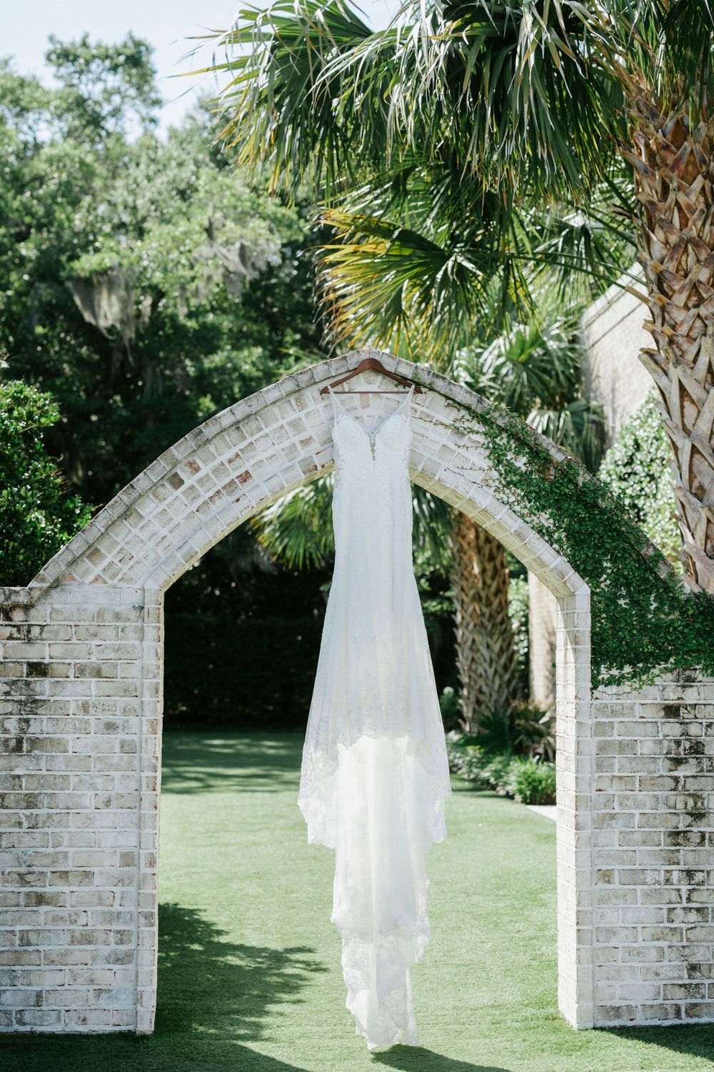 A white wedding dress hanging from a brick archway surrounded by lush greenery and palm trees in an outdoor garden setting.