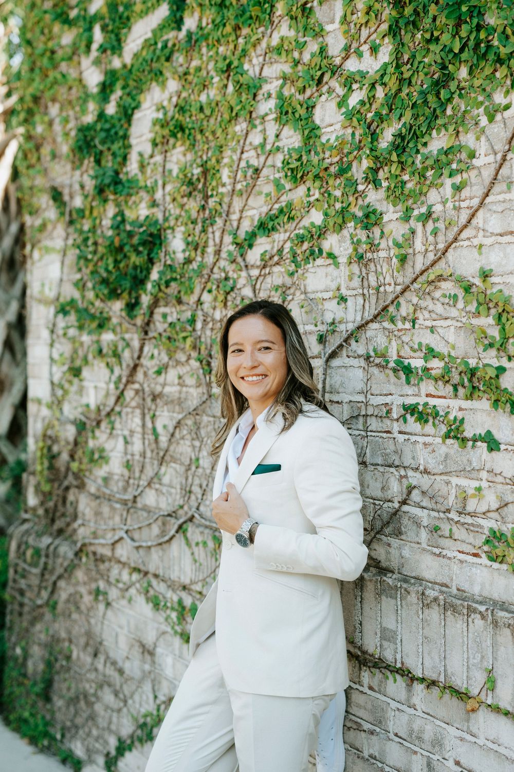 Woman in white suit smiling against ivy-covered brick wall outdoors.