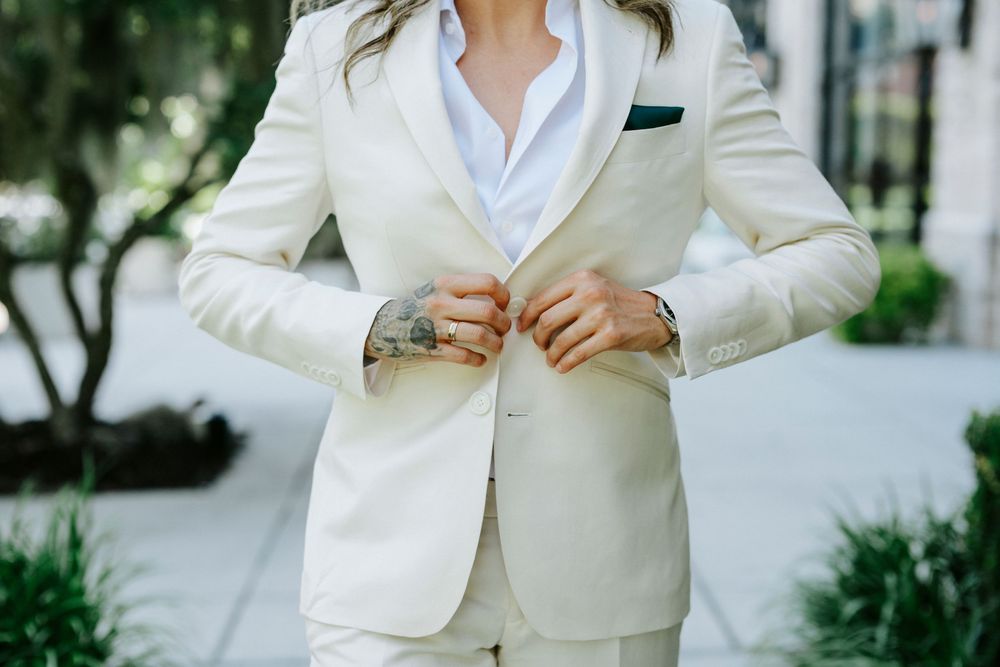 Woman in cream white pantsuit buttoning jacket outdoors, elegant bridal or formal attire with green pocket square.