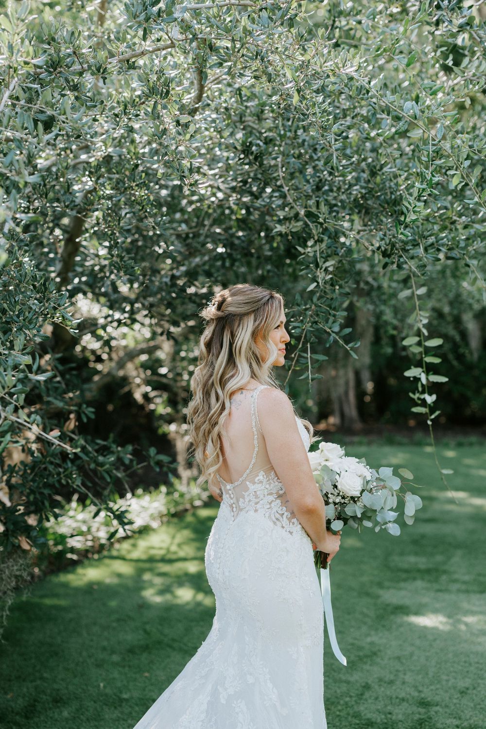 Bride in lace mermaid gown holding white bouquet stands under olive tree in lush garden.