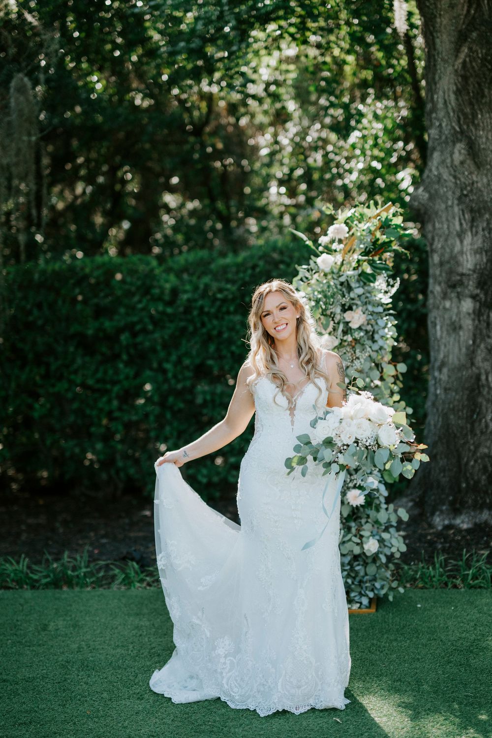 Smiling bride in lace wedding gown holding bouquet of white flowers beside floral arch in lush garden.
