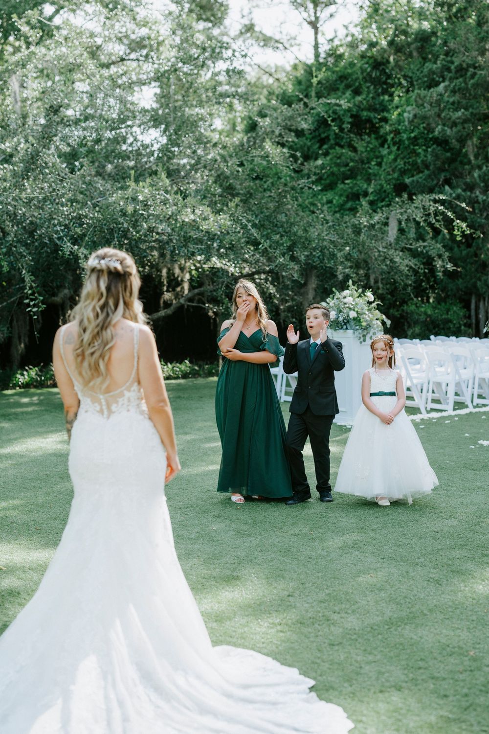 Bride in lace mermaid gown stands on green lawn as wedding party in emerald dresses reacts with joy outdoors.