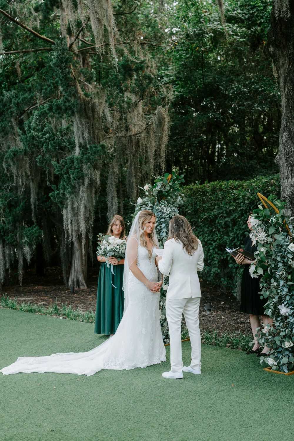Two brides exchange vows outdoors under a floral arch surrounded by lush greenery and Spanish moss-draped trees.