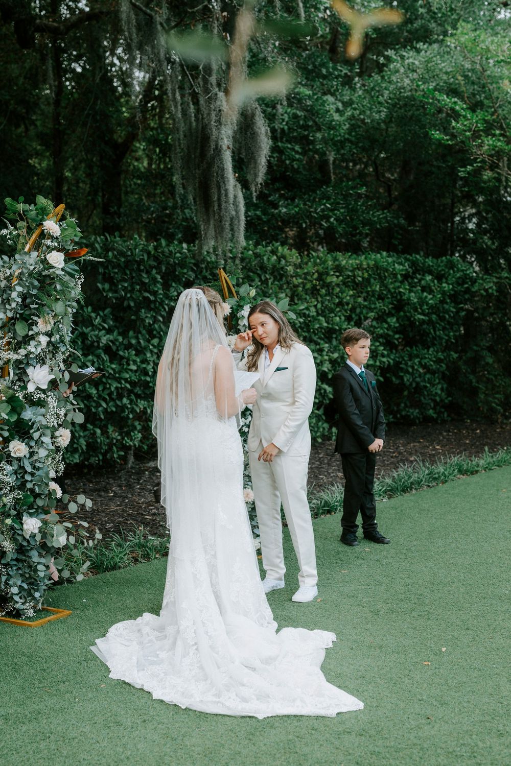 Outdoor wedding ceremony with bride in lace gown and veil exchanging vows with partner in white suit amid lush greenery.