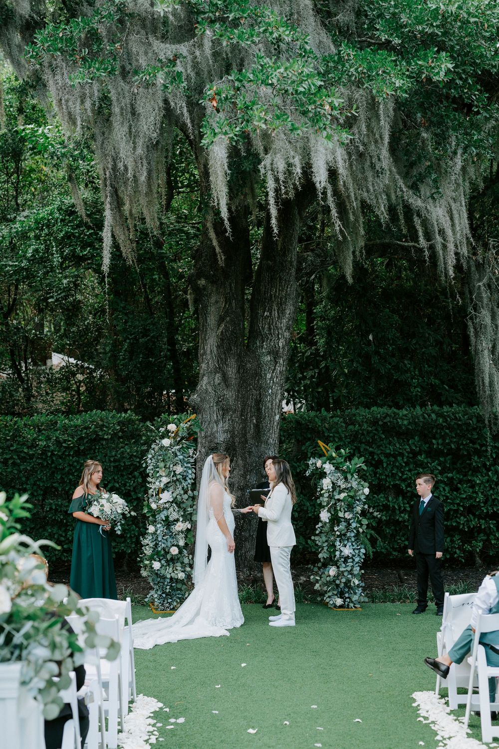 Two brides exchange vows under a moss-draped oak tree at an outdoor garden wedding ceremony.