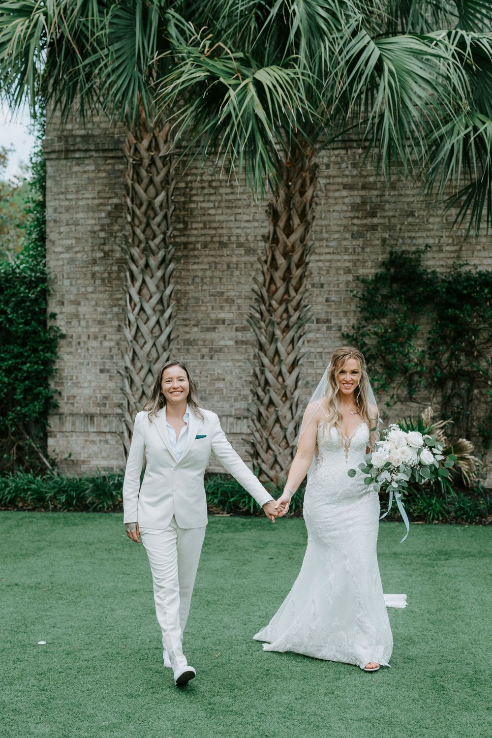 Two brides walk hand-in-hand on green lawn, one in white suit and one in lace gown, with palm trees backdrop.