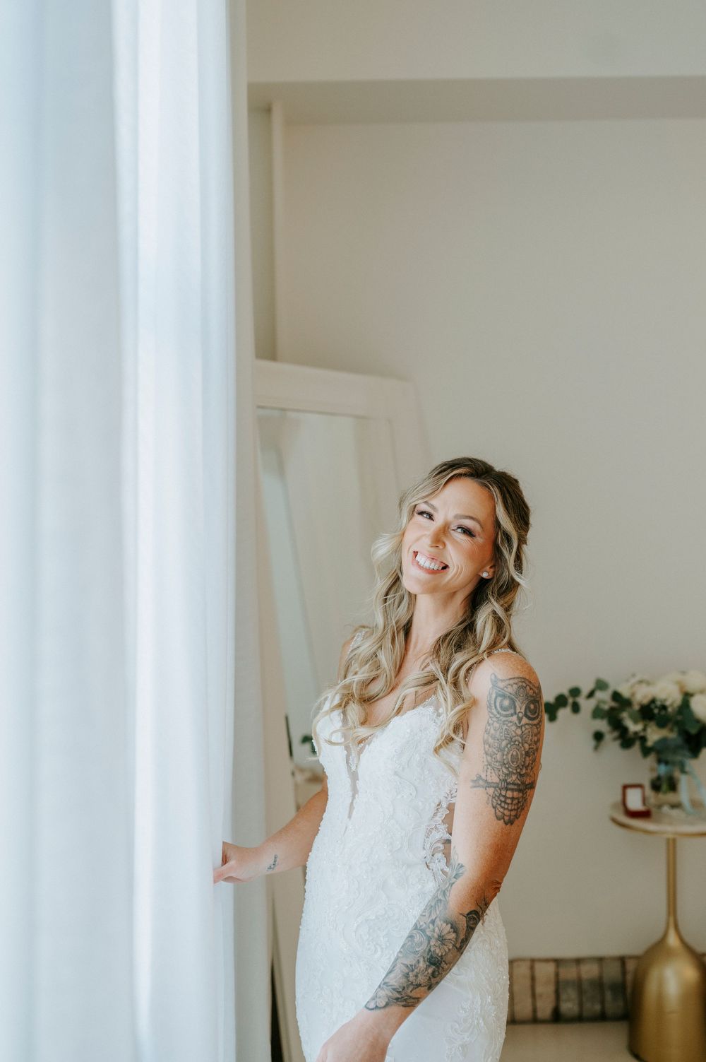 A smiling bride in a lace wedding gown with tattoos stands by a sheer curtain in soft natural light.