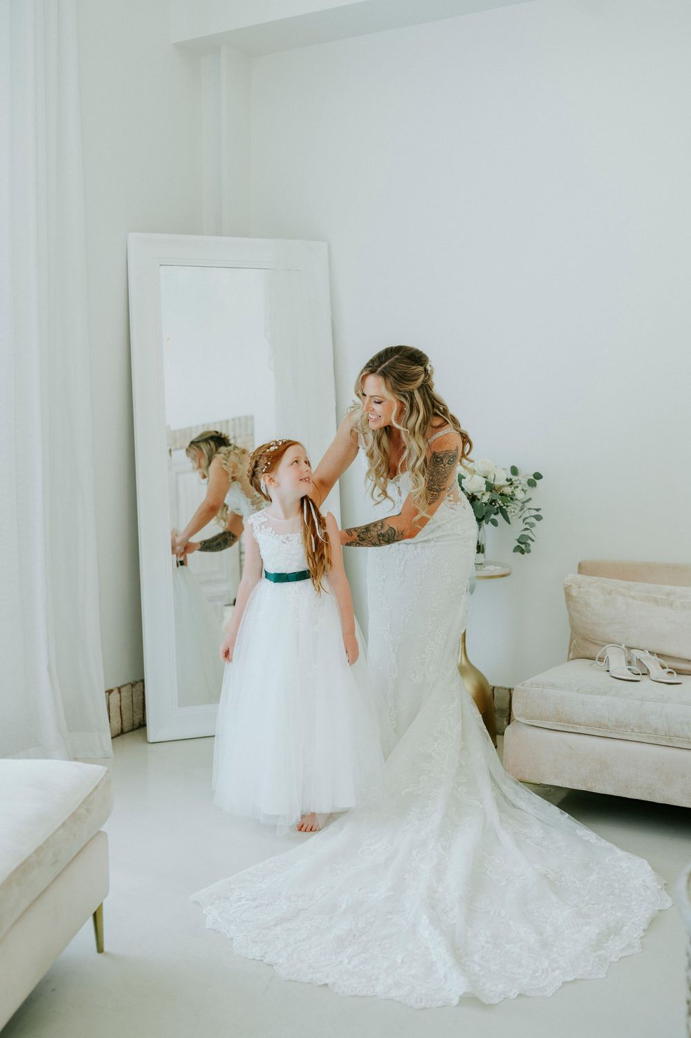 Bride in lace wedding gown adjusts flower girl's hair in bright white bridal suite with floor-length mirror.