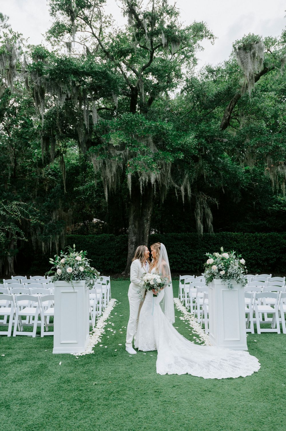 Two brides stand at outdoor altar flanked by white floral arrangements and chairs beneath Spanish moss-draped trees.