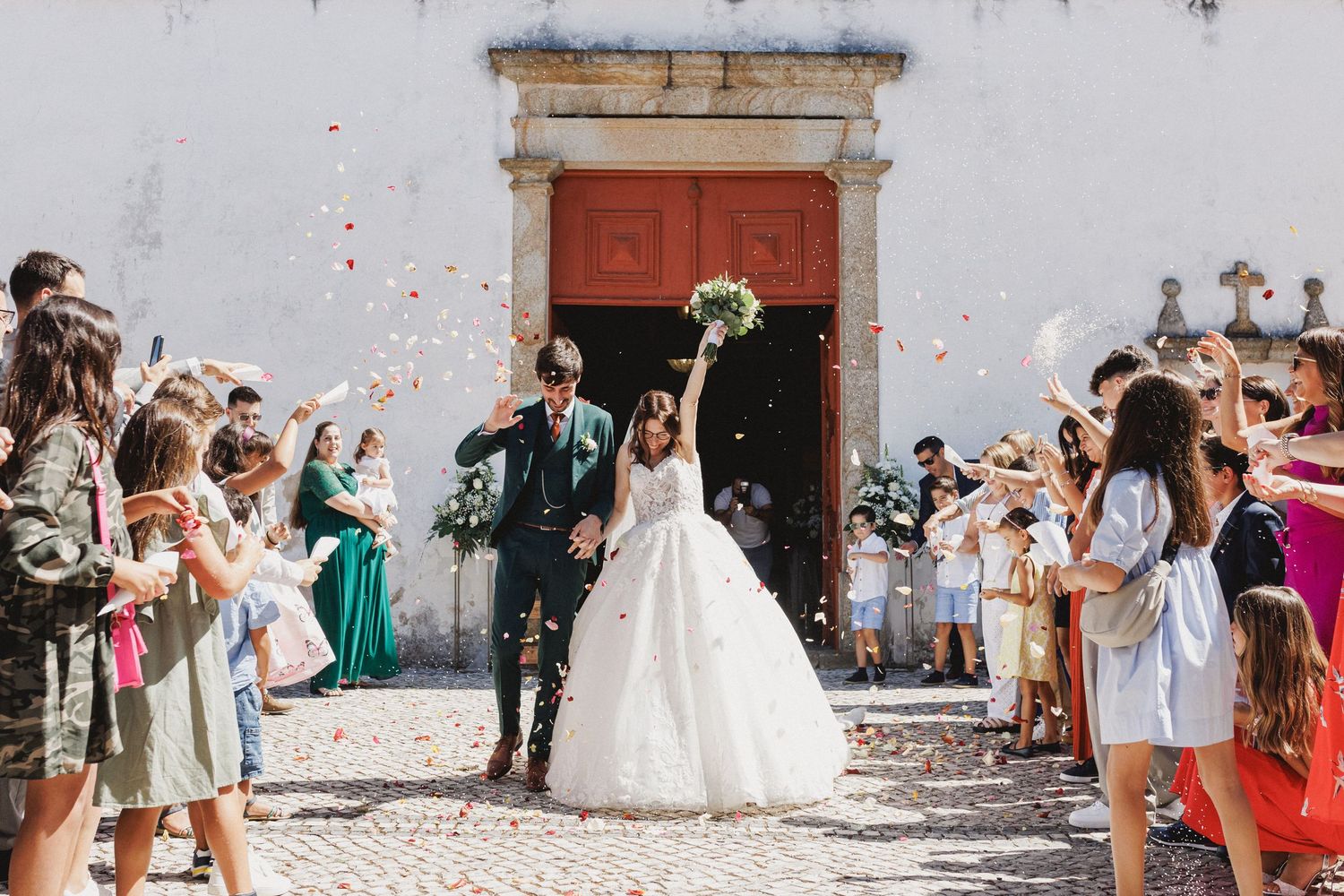 A joyful wedding celebration outside a church with confetti being thrown by guests as a couple exits through red doors.