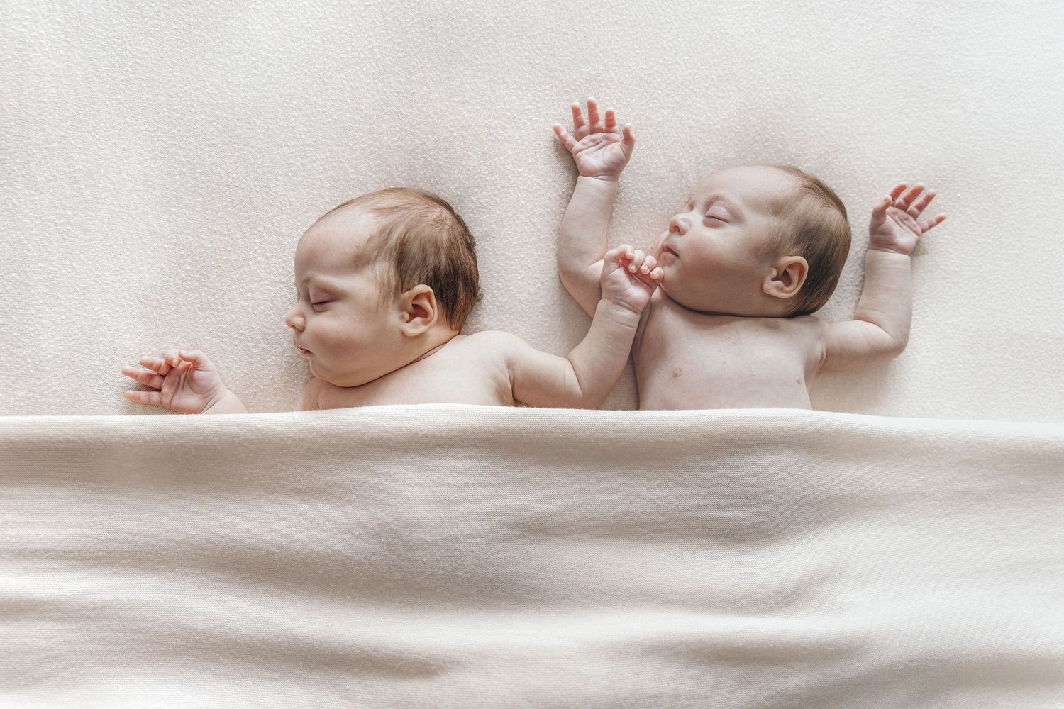 Two newborn twins lie side by side under a soft white blanket with arms raised in a peaceful pose.