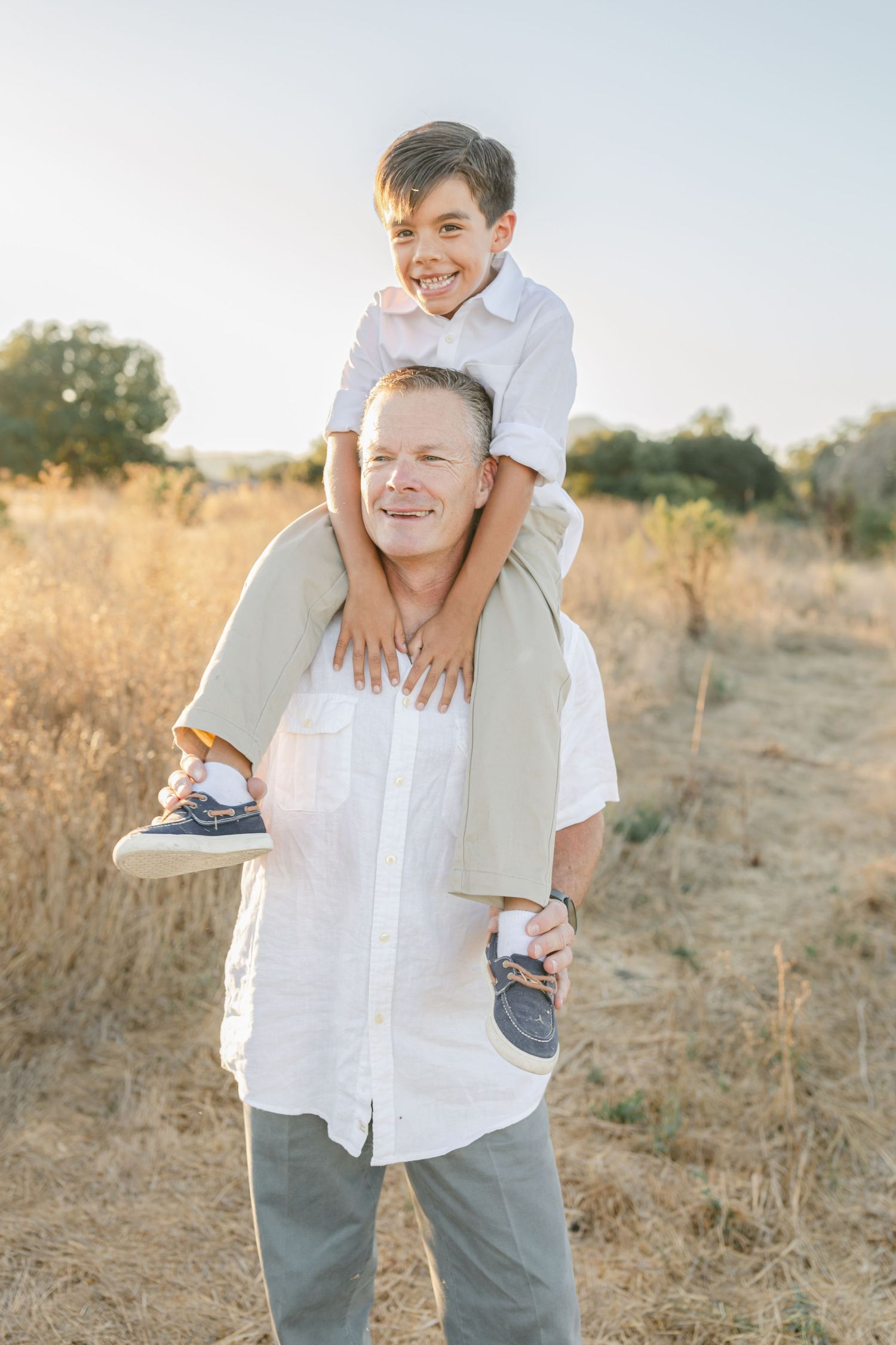 A smiling boy rides on a man's shoulders in a sunny field, both wearing white shirts outdoors.