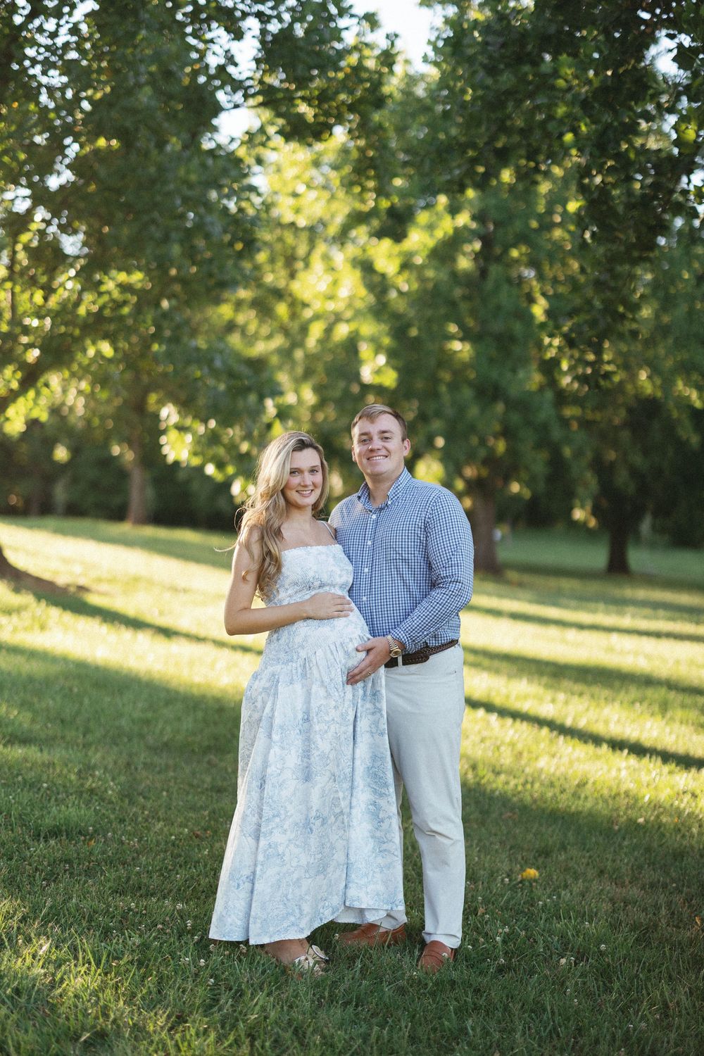 Smiling couple posing outdoors in a sunny park, woman in floral dress cradling baby bump, man in blue shirt beside her.