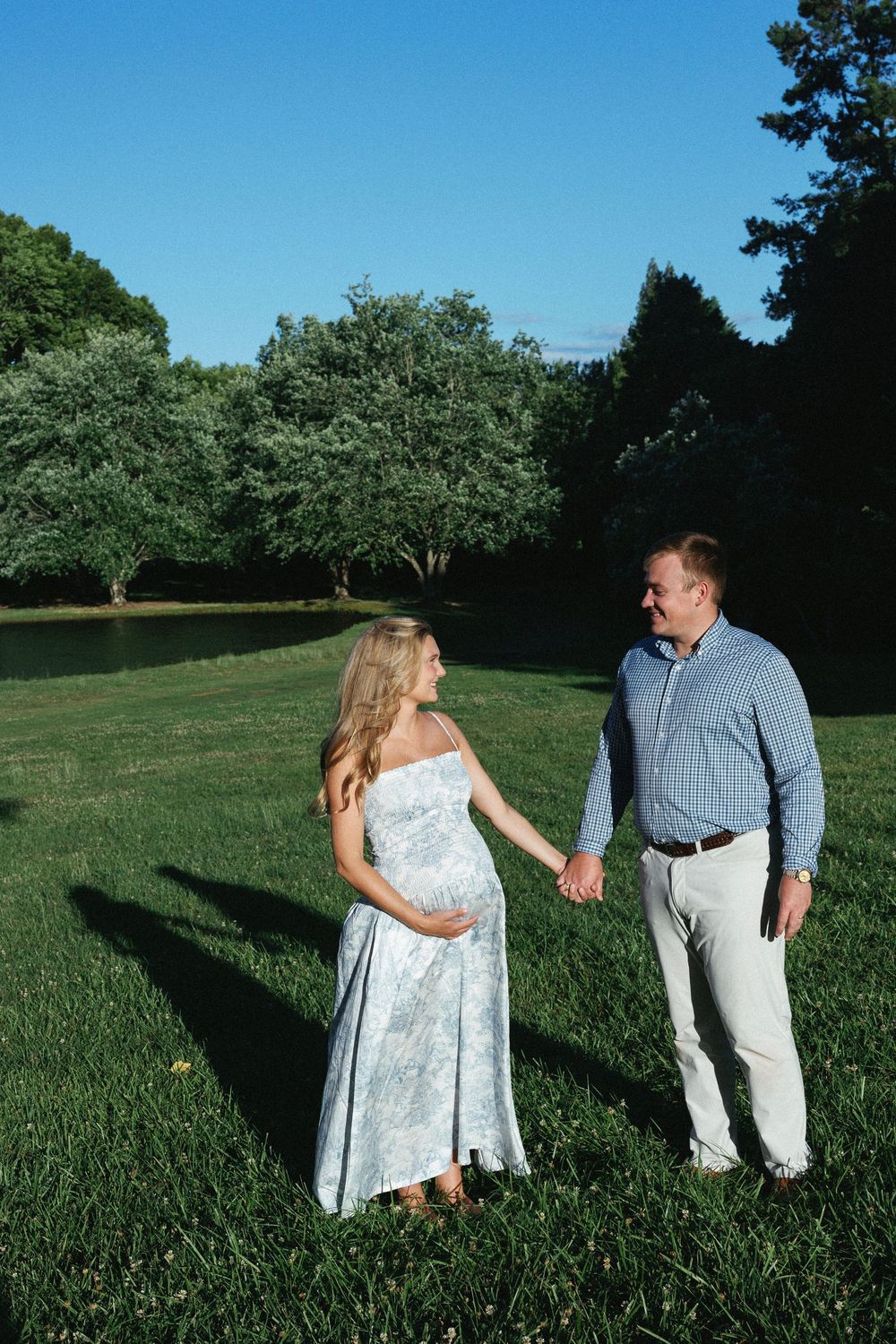 Pregnant woman in floral dress holds hands with man in blue shirt on green lawn surrounded by trees.