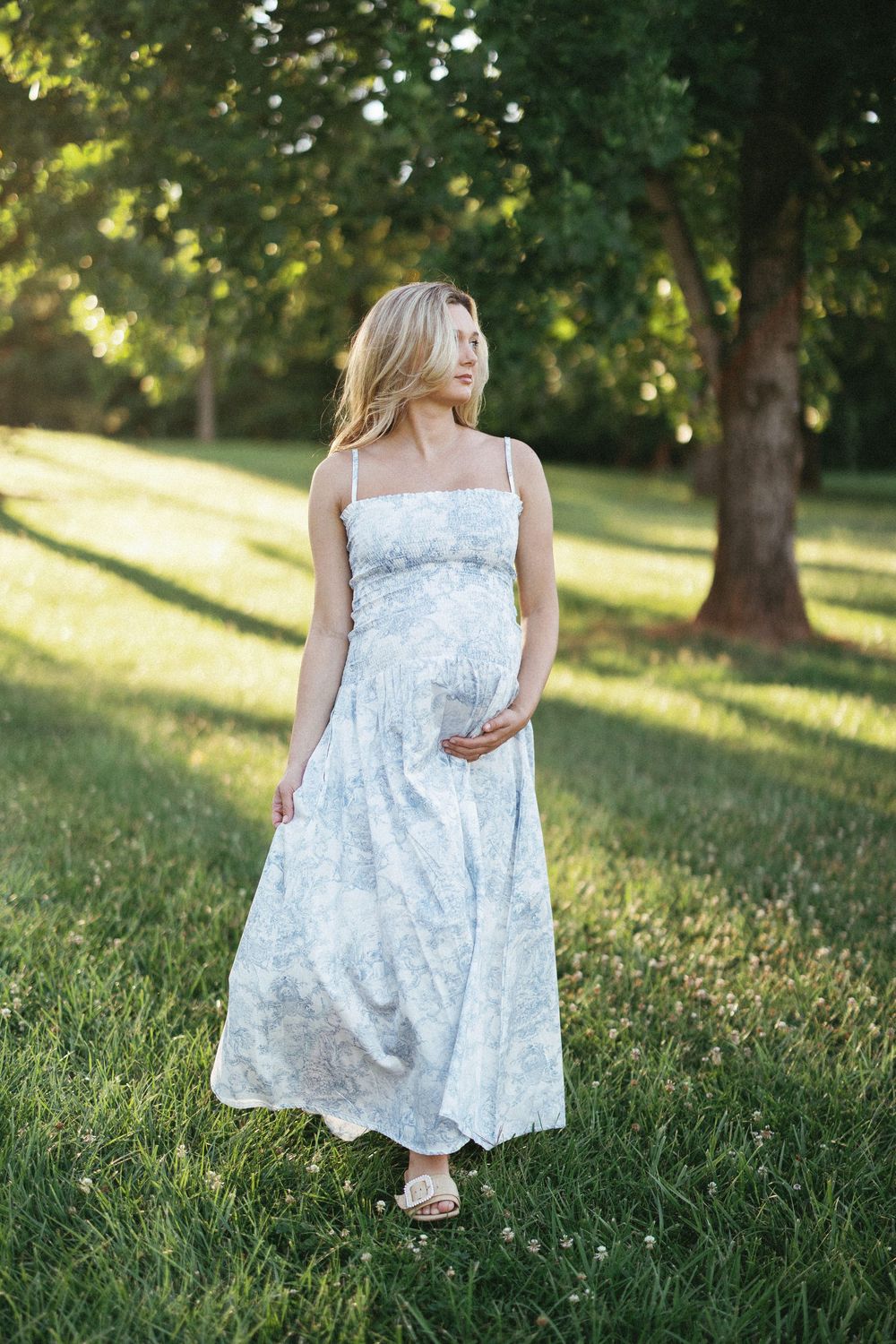 Pregnant woman in floral maxi dress standing in green park, cradling baby bump in golden sunlight.