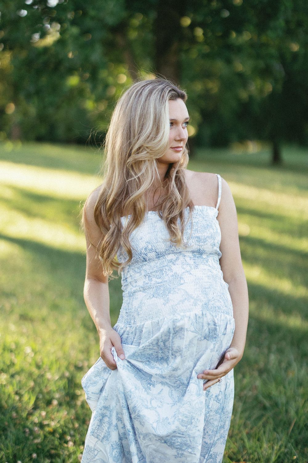 Pregnant woman in floral sundress standing in green park, long blonde hair, golden hour sunlight.