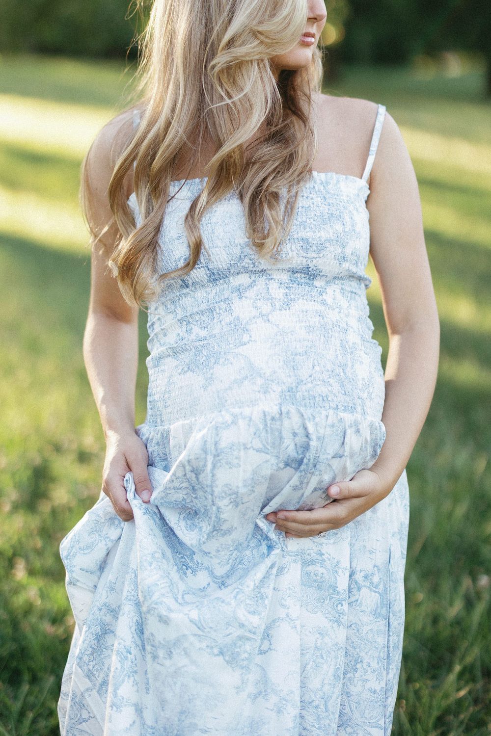 Pregnant woman in blue floral sundress holding baby bump outdoors in green field.