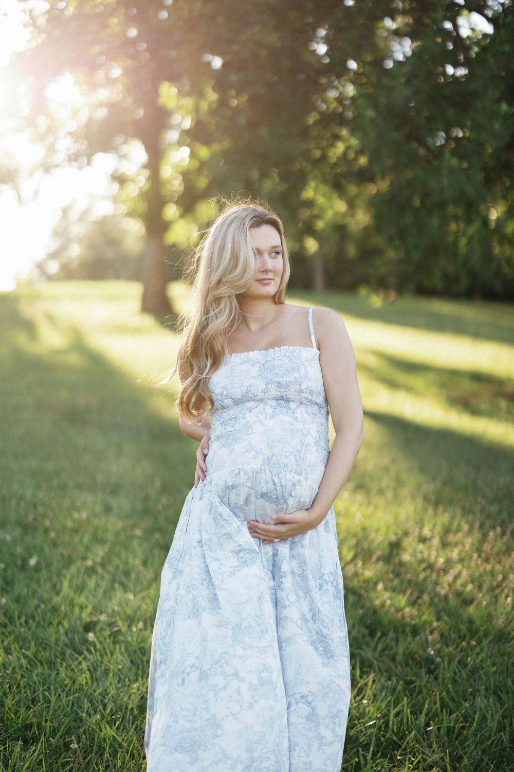 Pregnant woman in blue floral sundress cradling baby bump outdoors in sunlit green park.