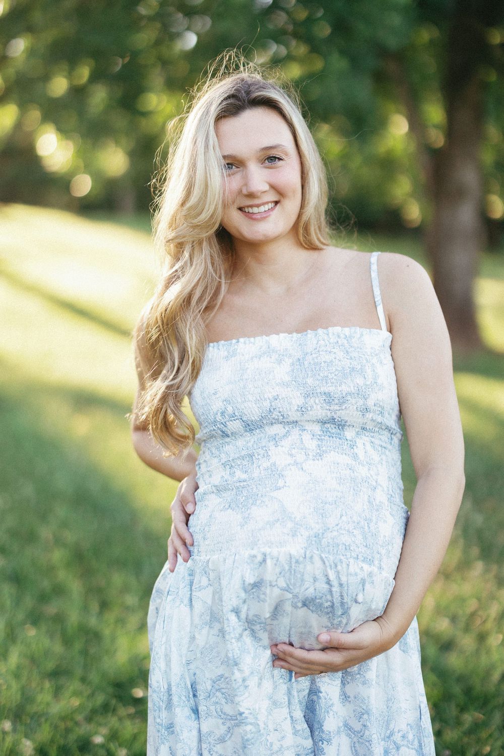 Smiling pregnant woman in blue floral sundress cradling baby bump outdoors in sunny green park.