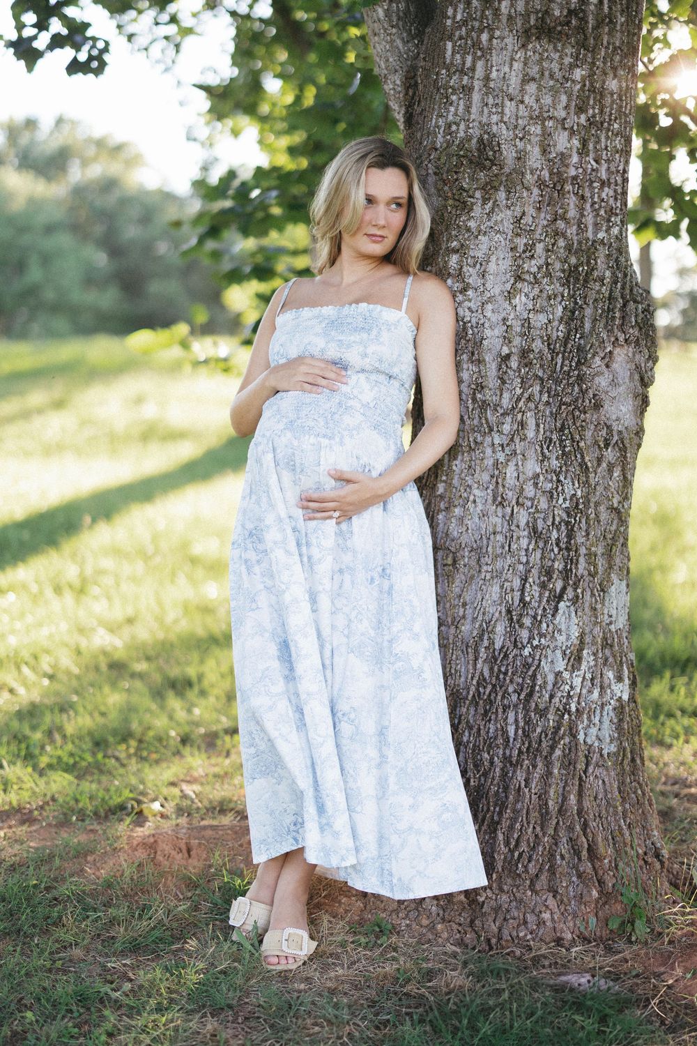 Pregnant woman in light blue floral maxi dress leaning against tree in sunny park.