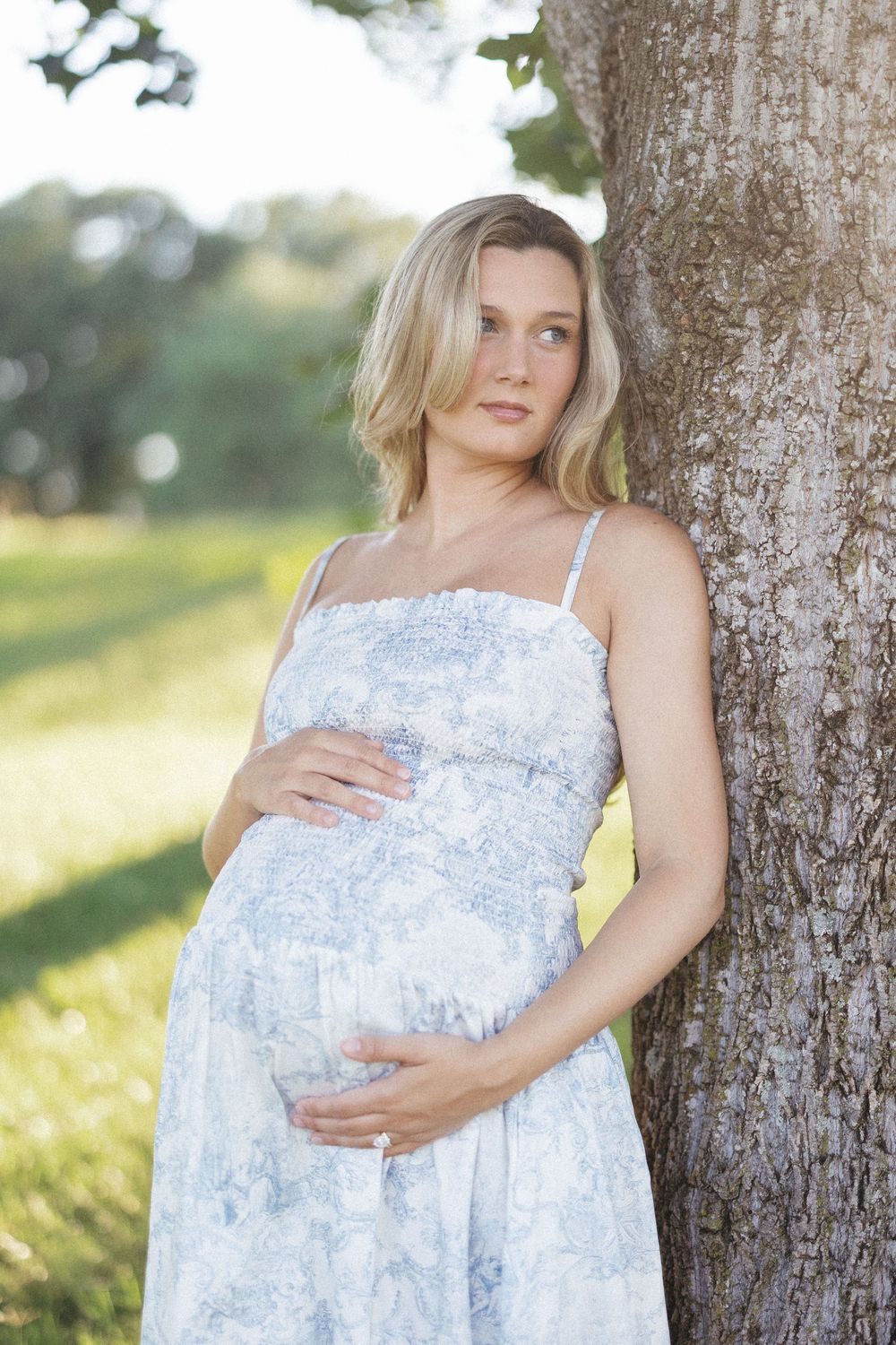 Pregnant woman in blue floral dress leaning against tree, cradling baby bump outdoors in sunny park.