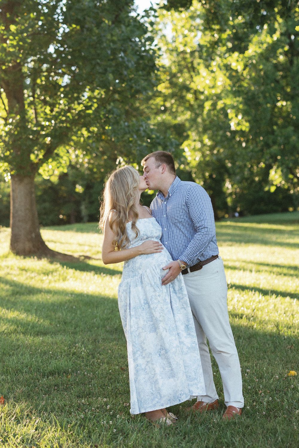 Couple sharing a kiss in a sunlit park, woman in floral maternity dress cradling baby bump.
