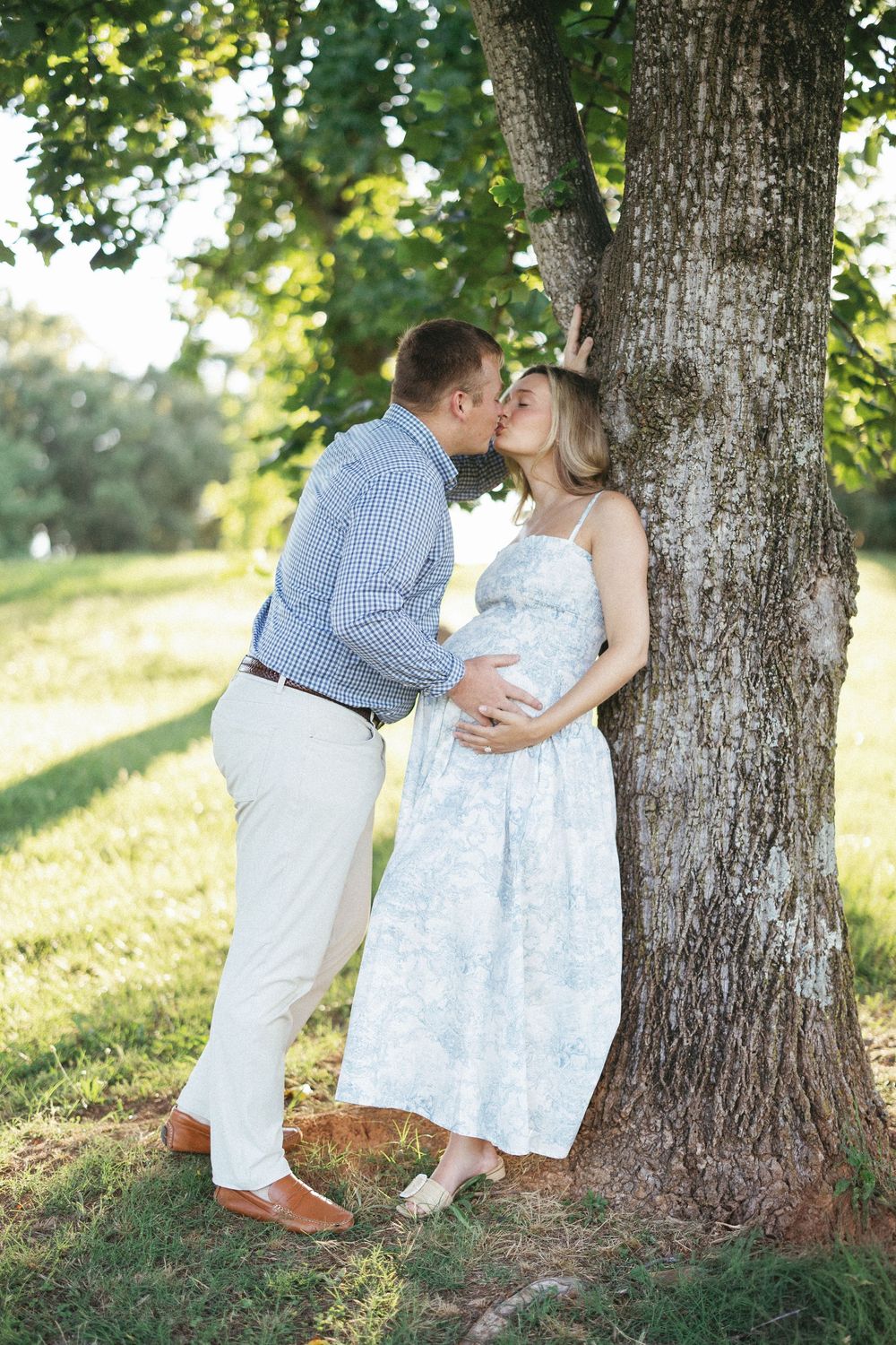 Pregnant couple sharing a kiss beside a large tree in a sunny green park, woman in floral dress cradling her baby bump.