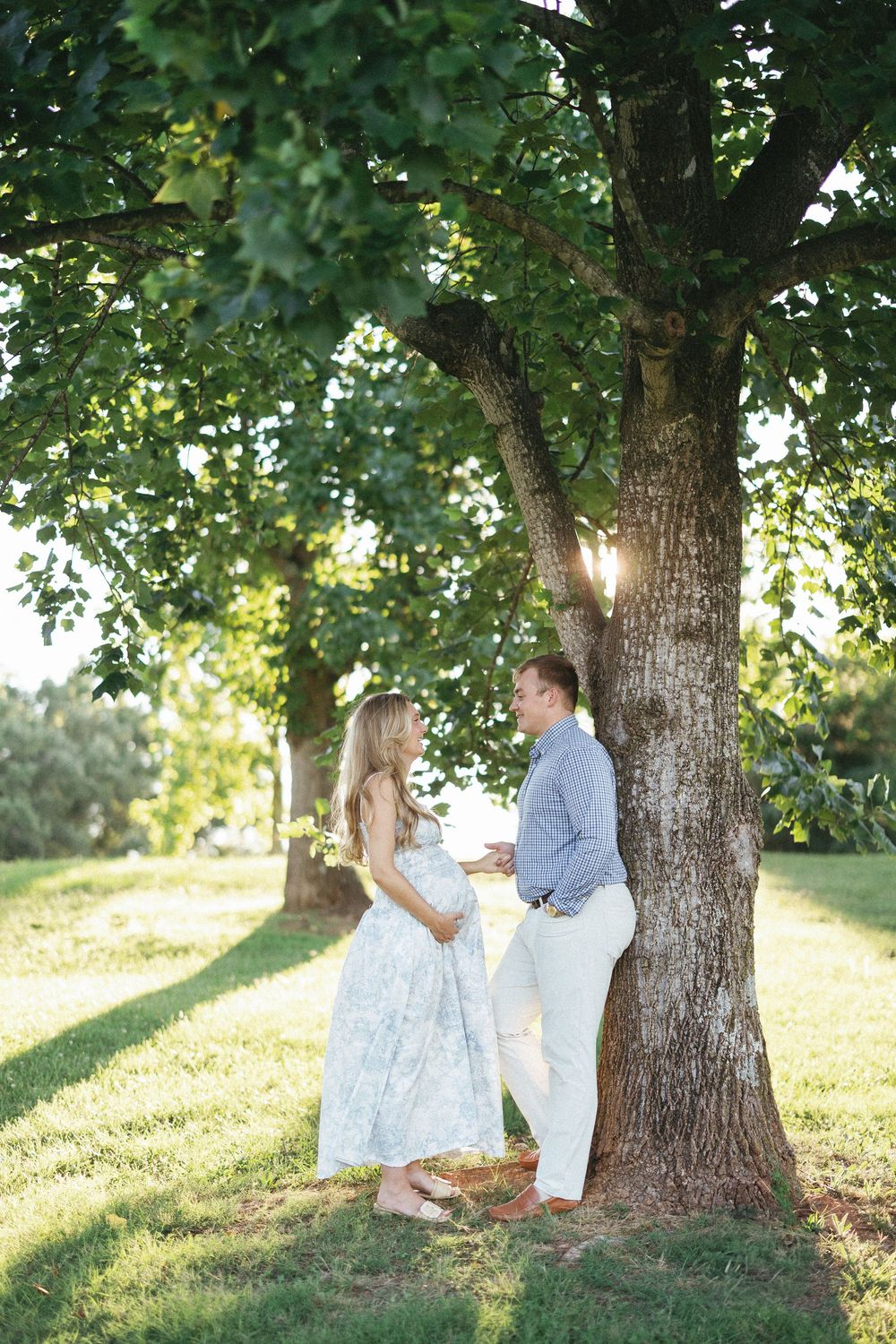 Pregnant couple sharing a tender moment under a large tree in a sunny green park, woman in floral dress.